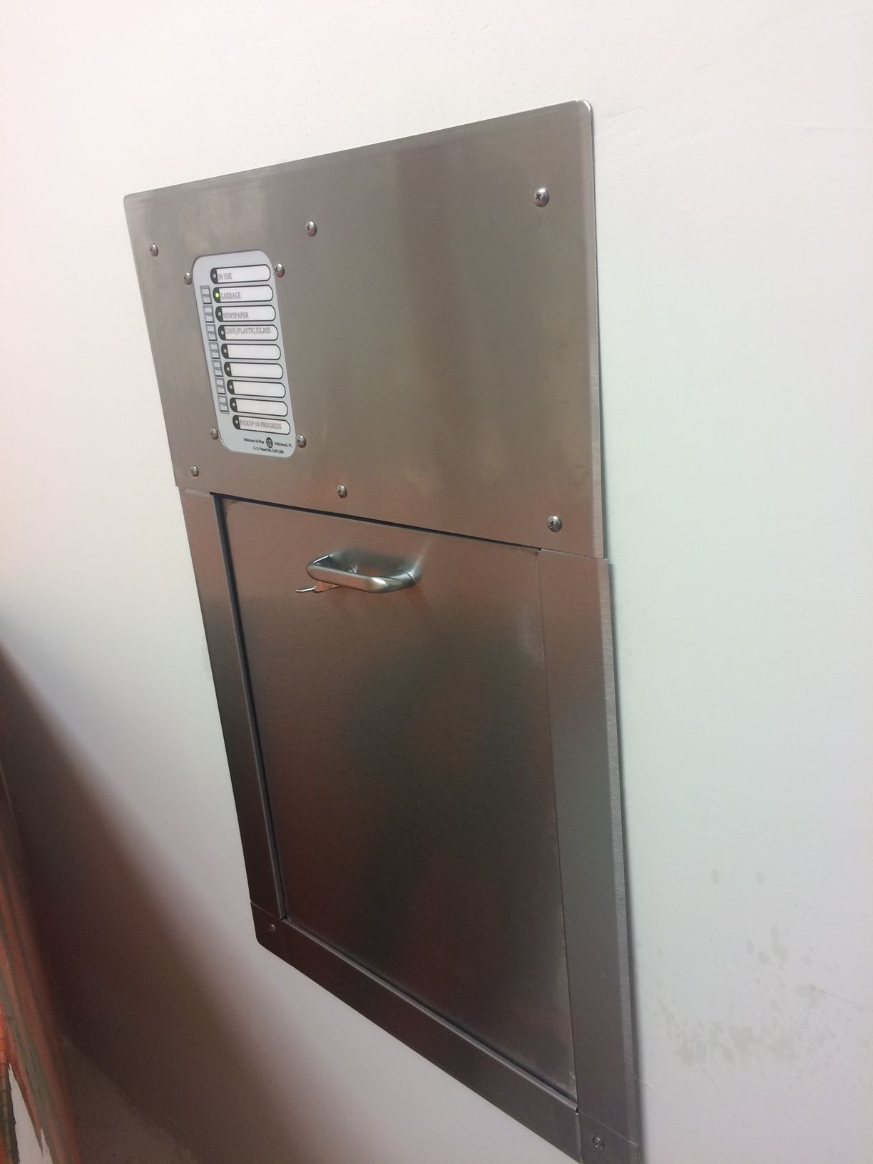 Stainless steel wall-mounted trash receptacle with hinged door and small label strip on the upper section.