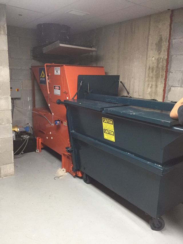 An industrial trash compactor and a dumpster inside a concrete room. Orange compactor, dark dumpster.