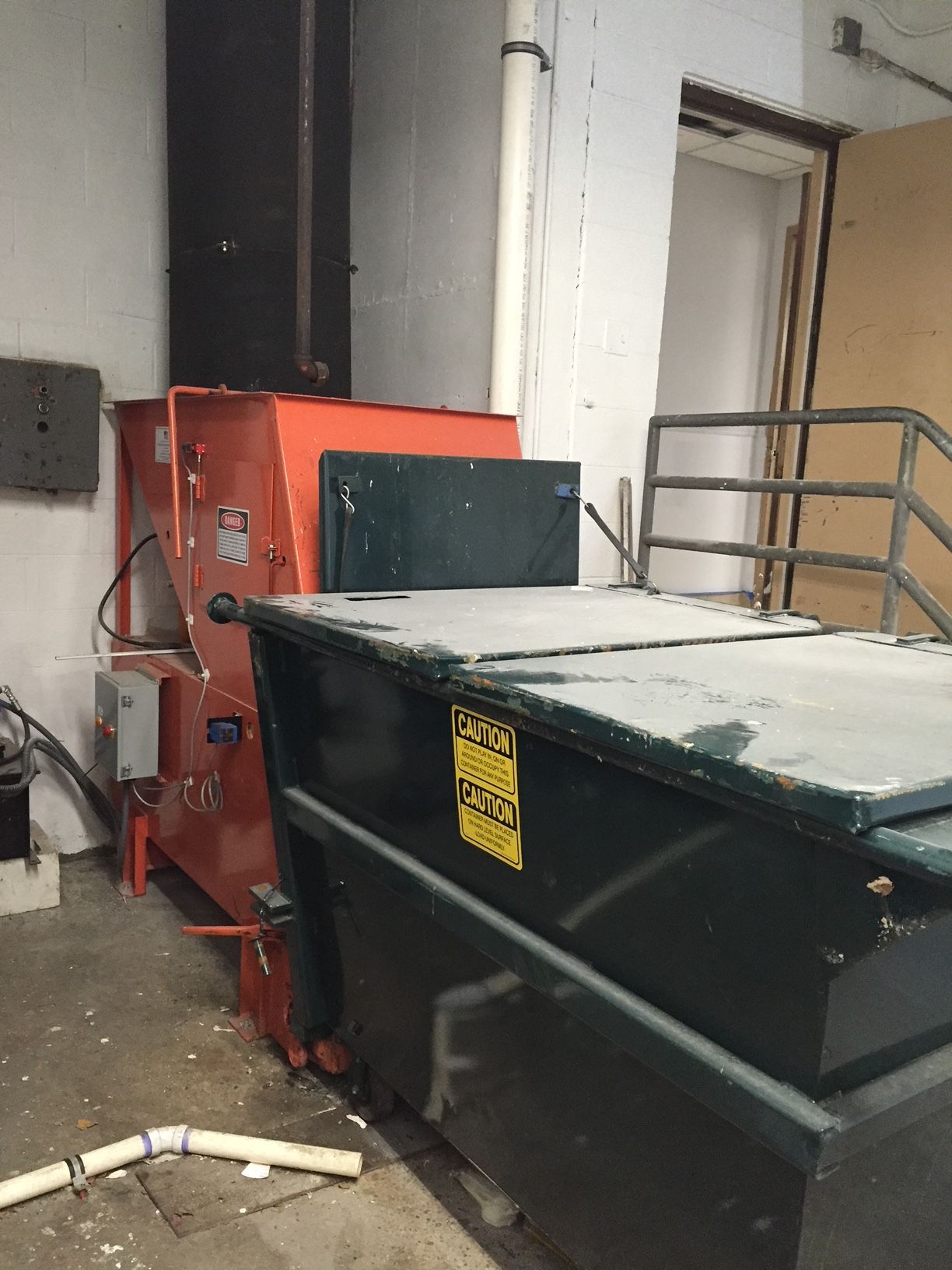 Orange trash compactor next to a large dumpster in an indoor setting.