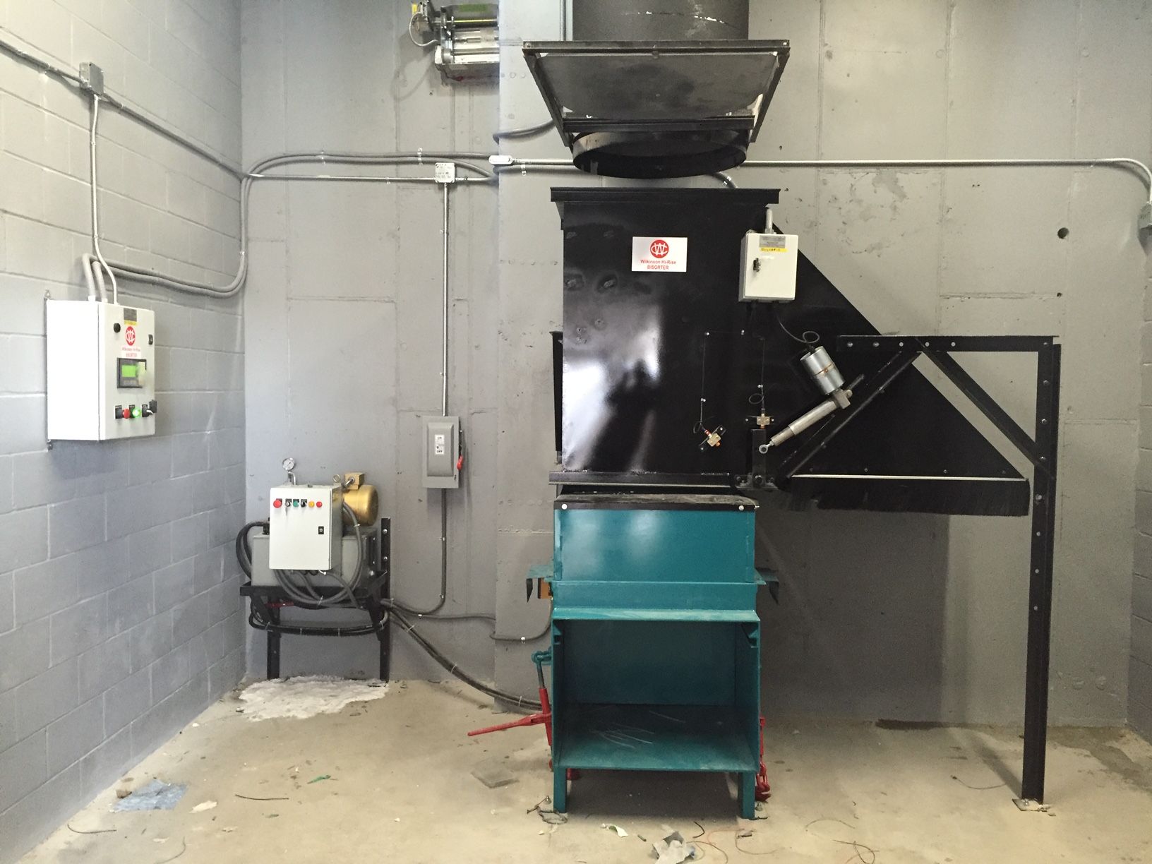 A black and teal industrial trash compactor in a gray-walled room.