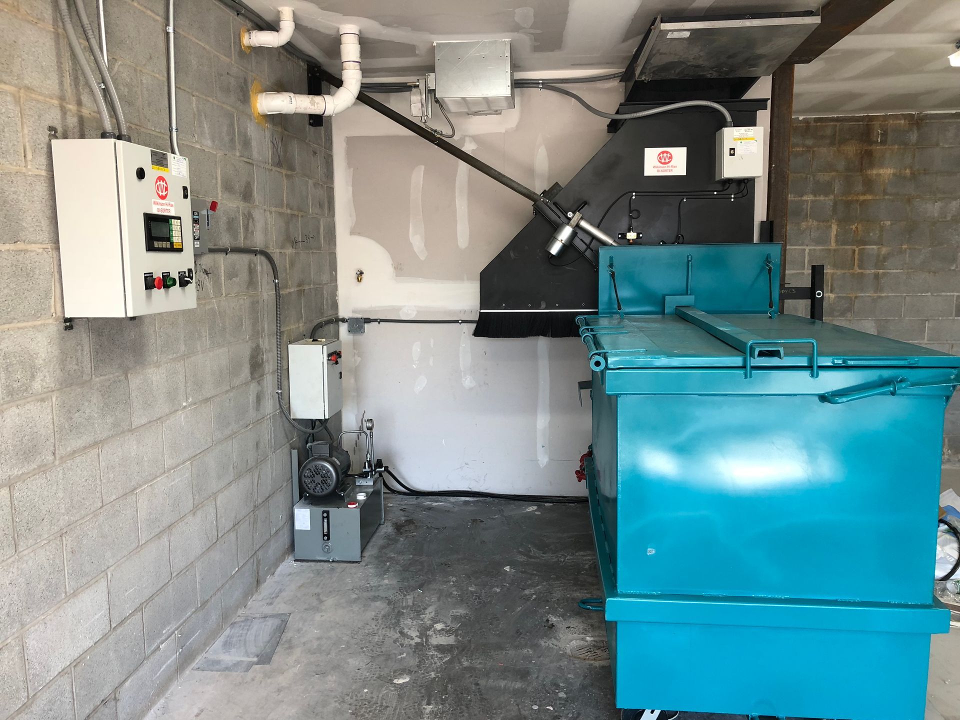 Blue industrial machine in a cinder block room with control panel.