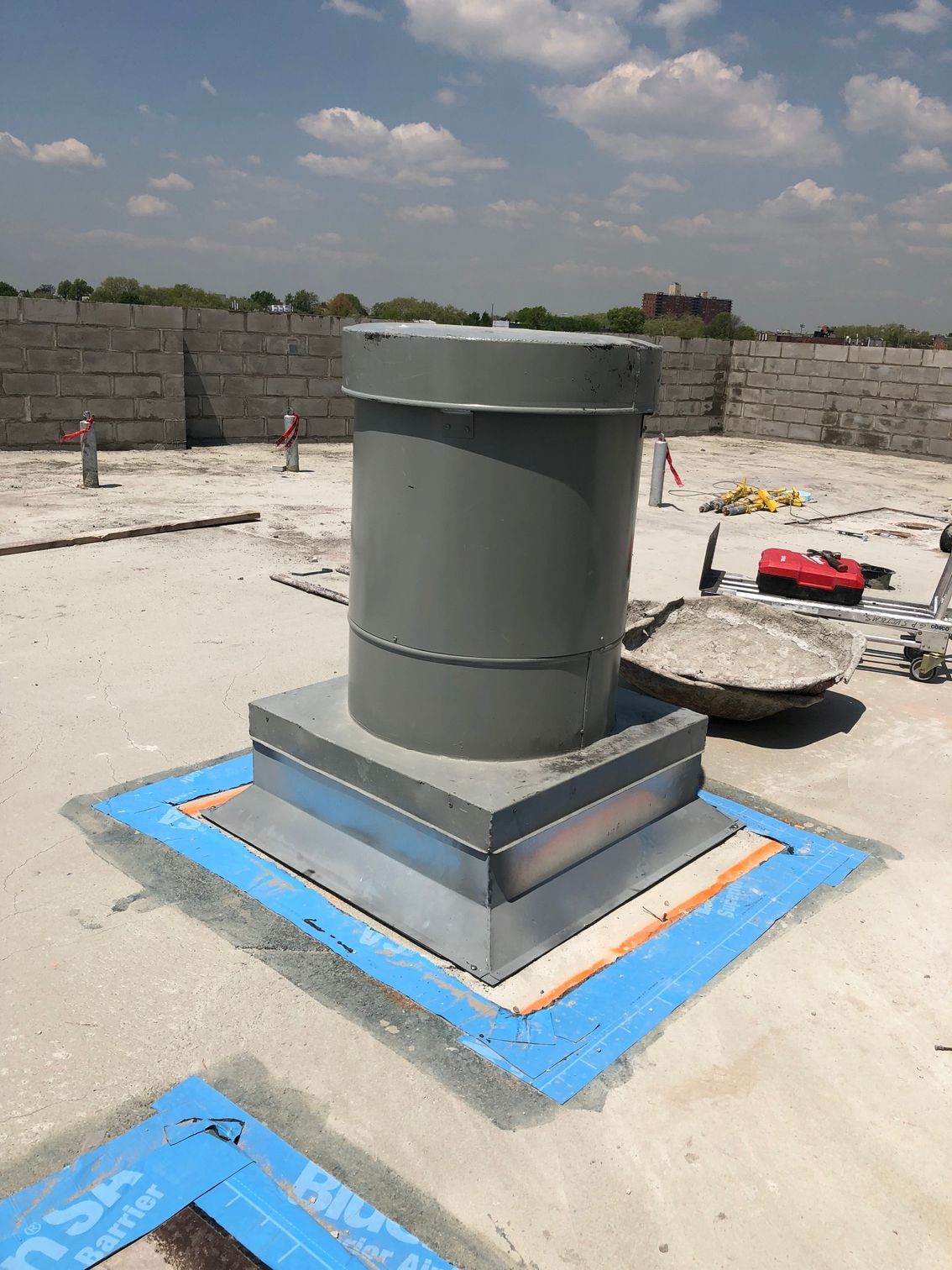 Gray ventilation pipe on a rooftop, sealed with blue and orange material. Construction in progress, sunny day.