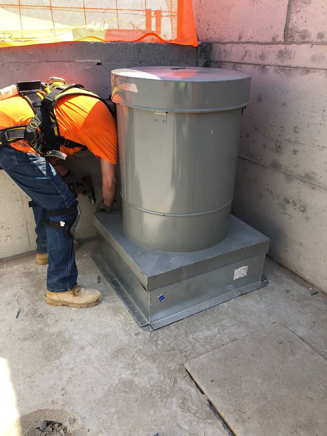 Worker in orange shirt and safety gear installing large gray cylindrical electrical equipment on a concrete surface.