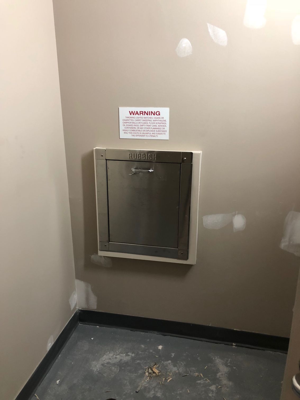 A stainless steel access panel on a tan wall with a warning sign above. A dark floor is below.