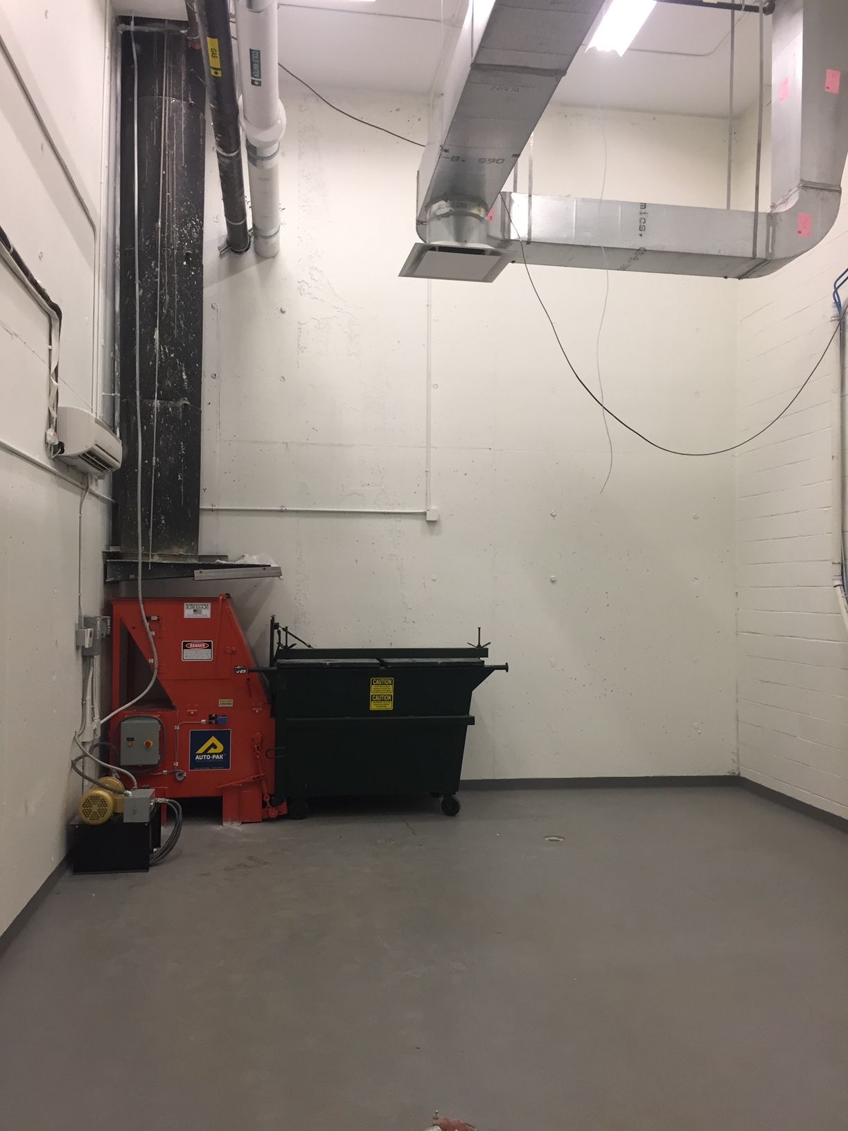 A room with trash bin, shredder, ducts, and pipes against white walls and a gray floor.