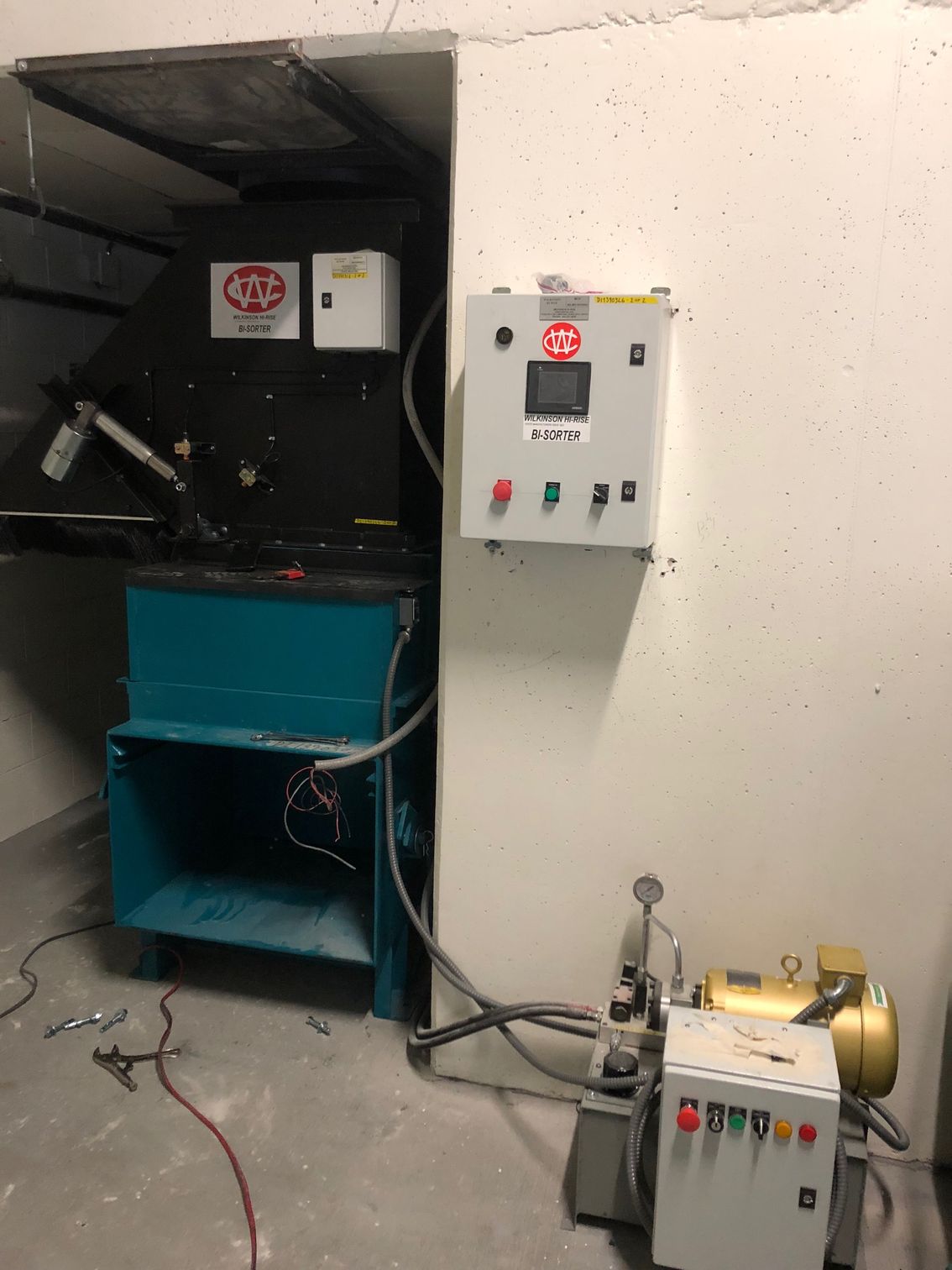 Industrial equipment in a corner, with a black machine, teal base, and control panels on the wall.