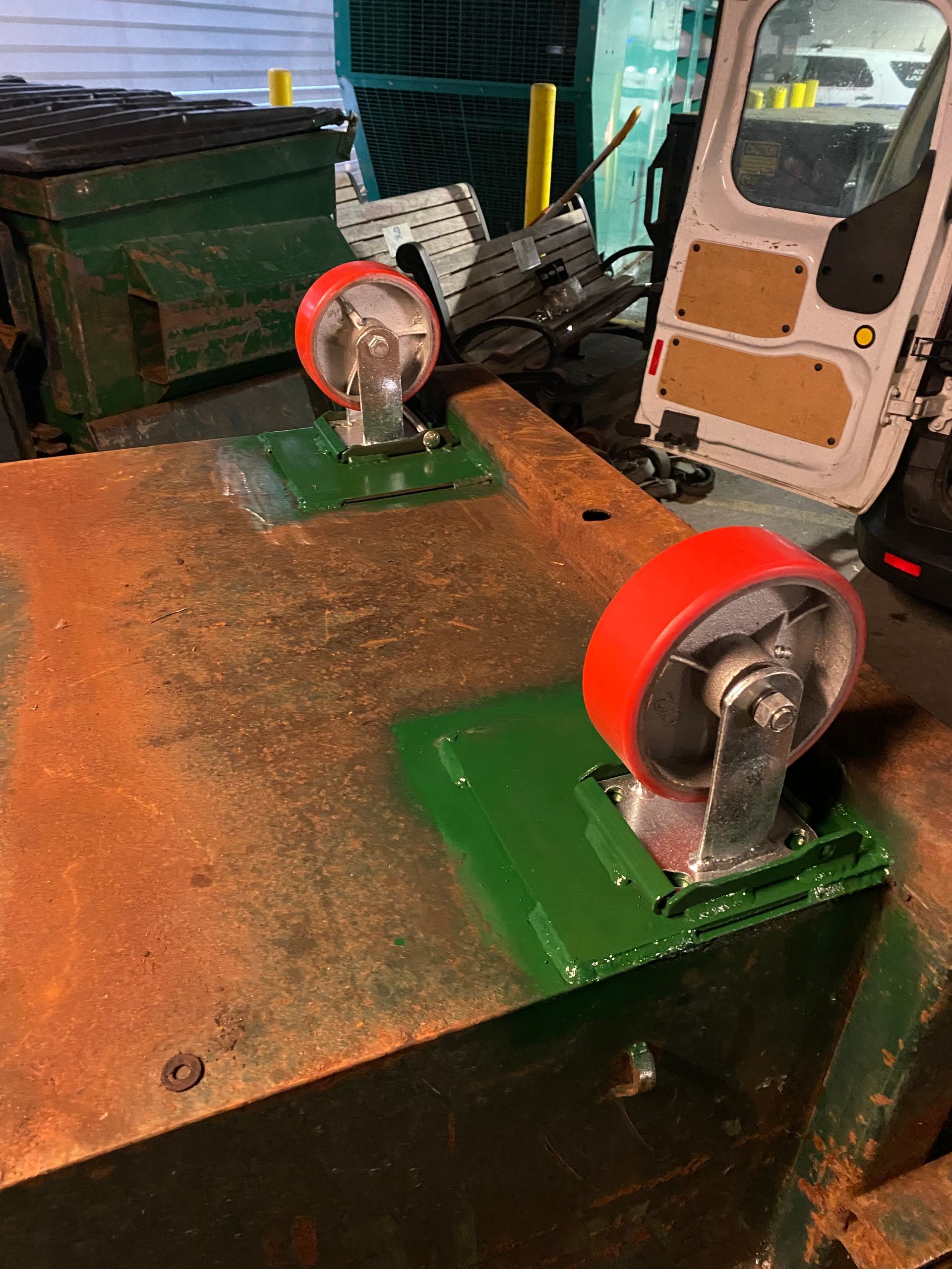Two red-wheeled casters mounted on green painted metal plates on a rusty brown surface.