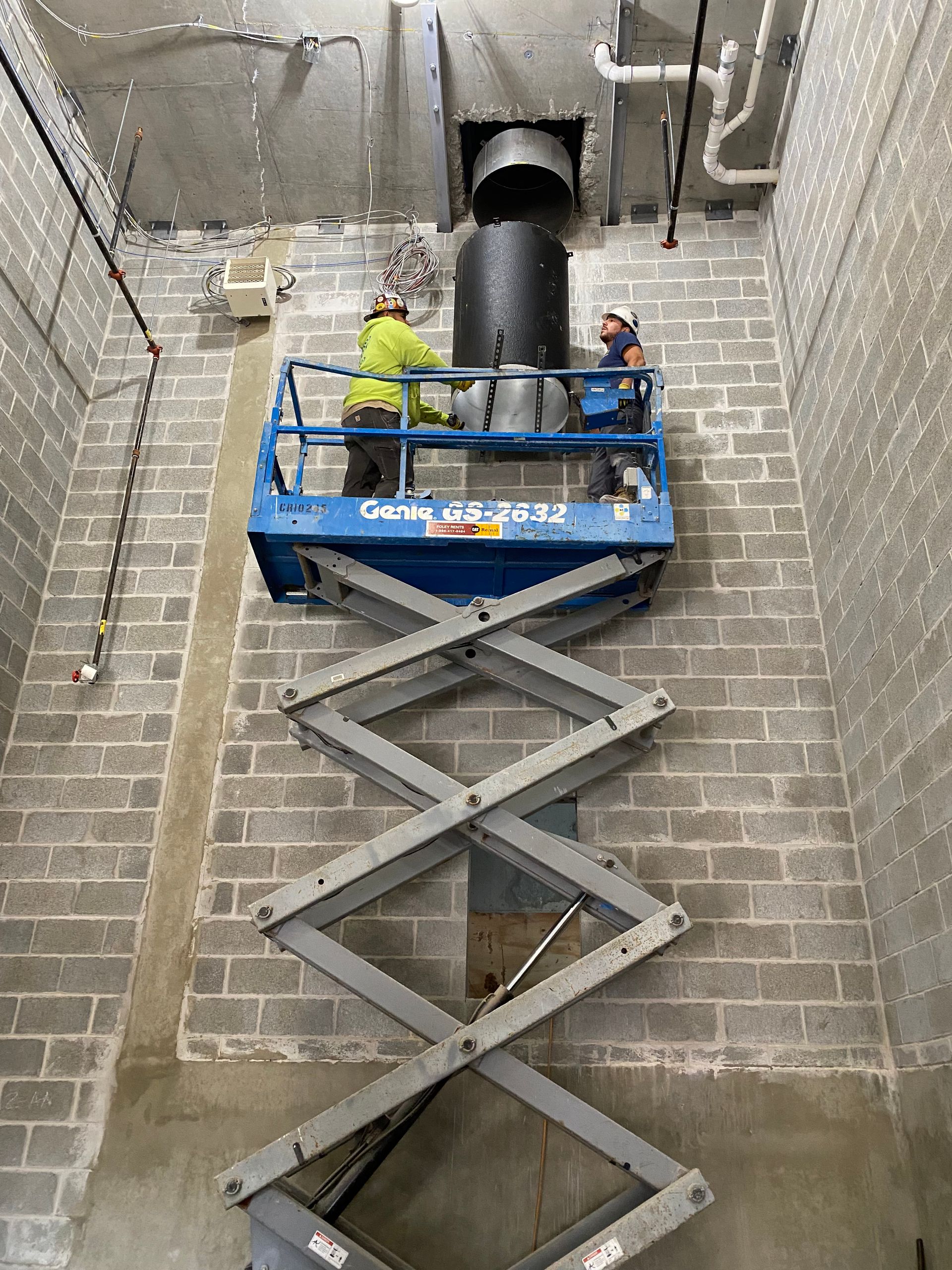 Two workers on a blue lift installing ductwork in a concrete block wall building.