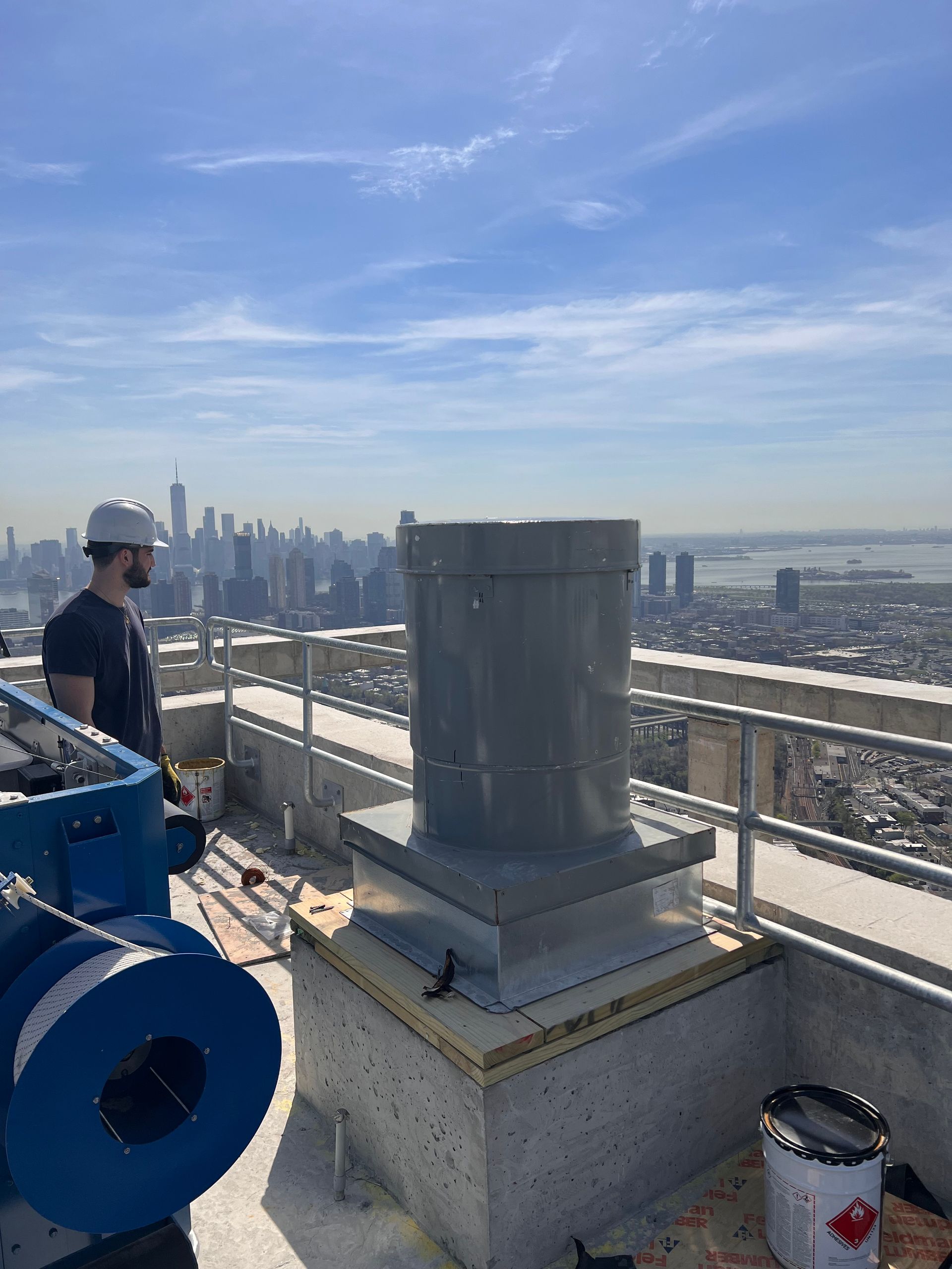 Man in hard hat looks out at city skyline from rooftop near ventilation equipment.