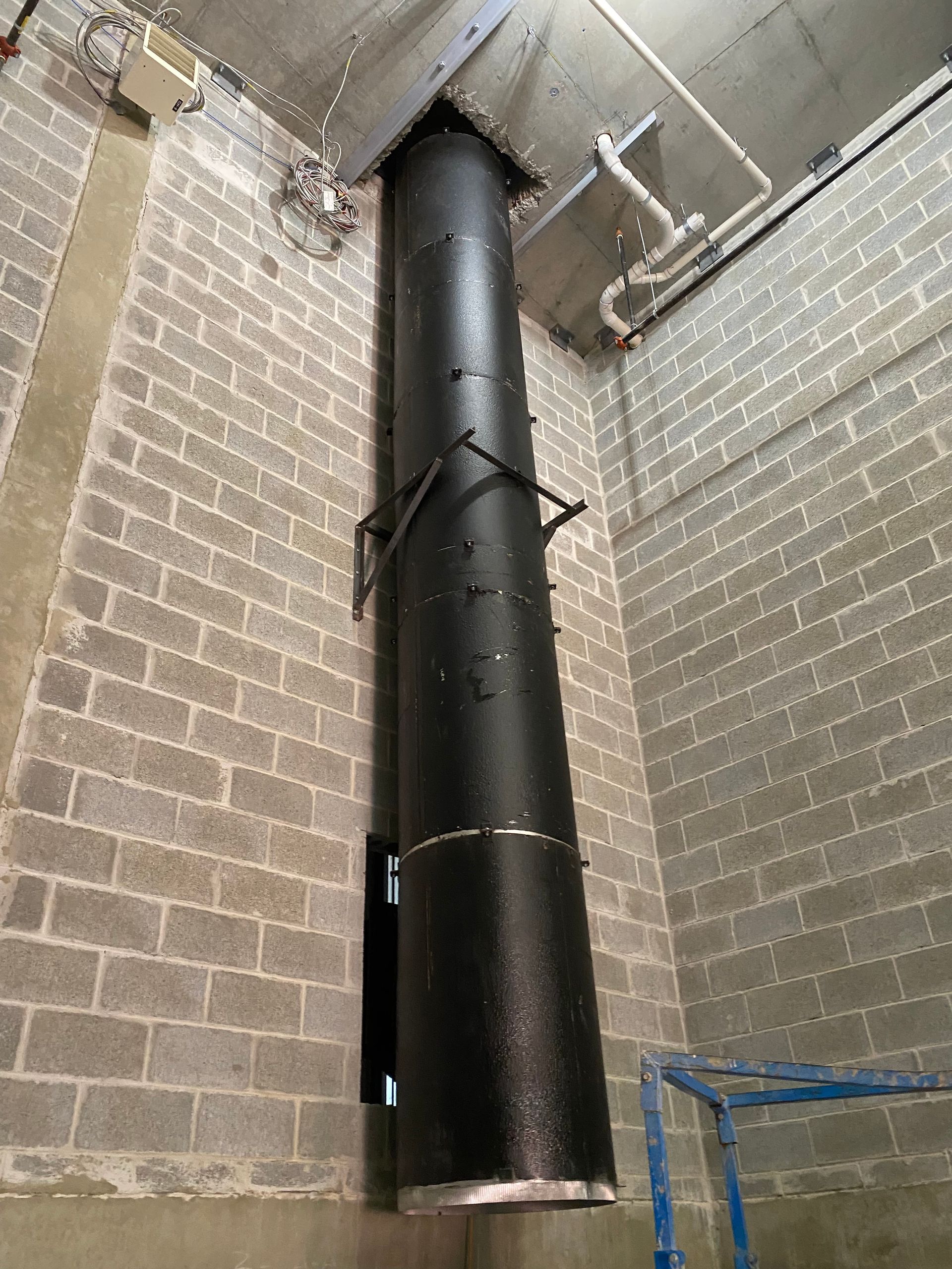 Black cylindrical vent pipe mounted on a gray cinder block wall. Blue scaffolding visible.