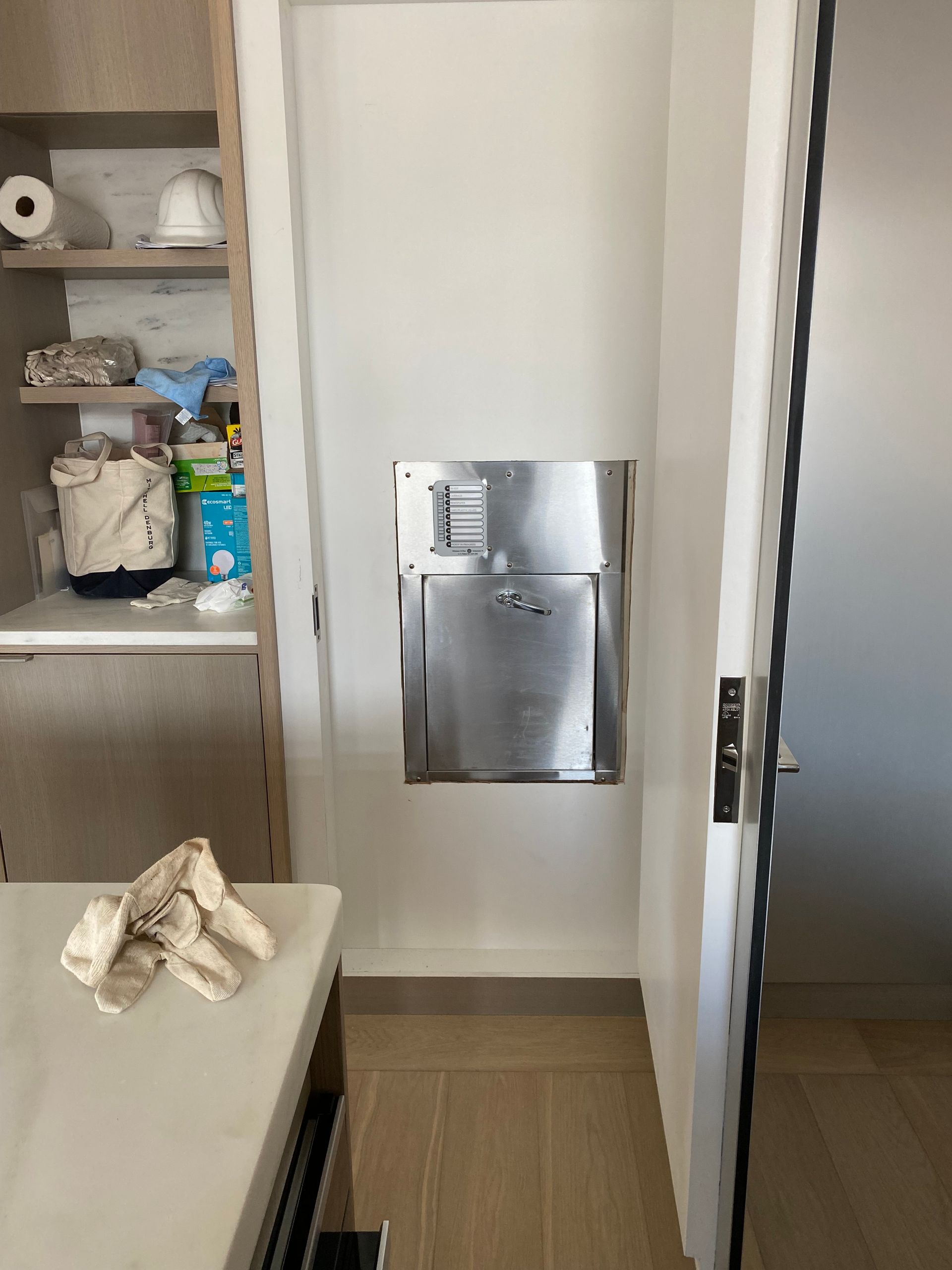 Stainless steel recessed trash receptacle on white wall in a narrow hallway. Door on the right, cabinet on the left.