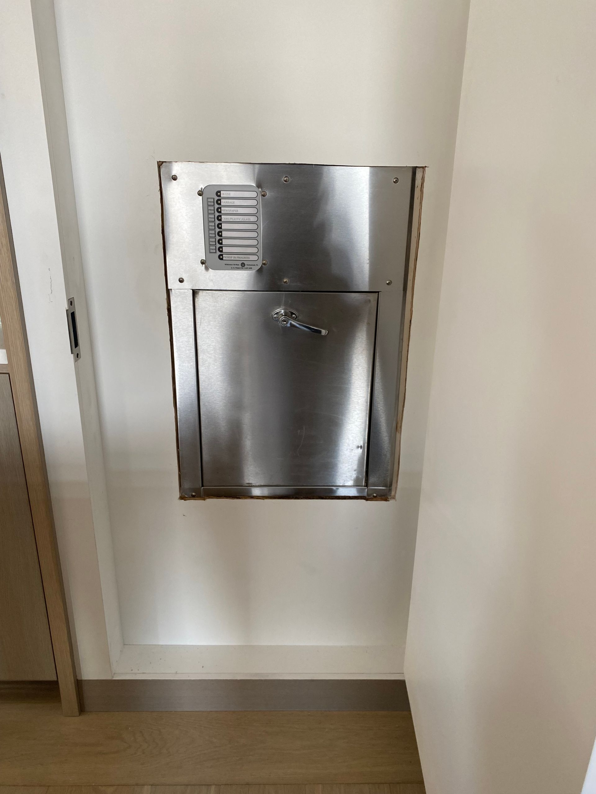 Stainless steel wall-mounted trash chute with a rectangular opening and air vent. Mounted on a white wall.