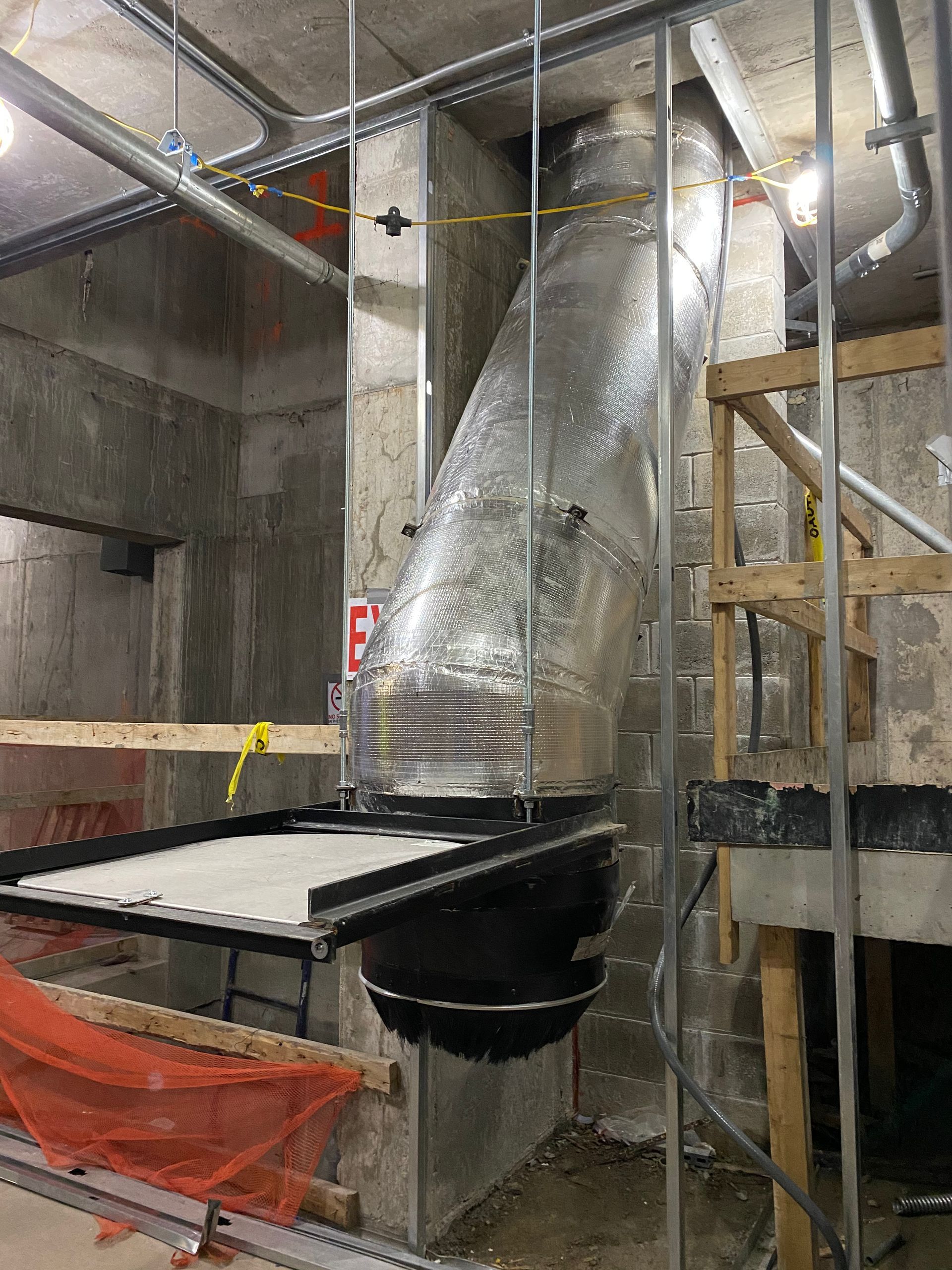 Construction site with HVAC ductwork, silver, black, and concrete walls. A temporary wooden platform is visible.