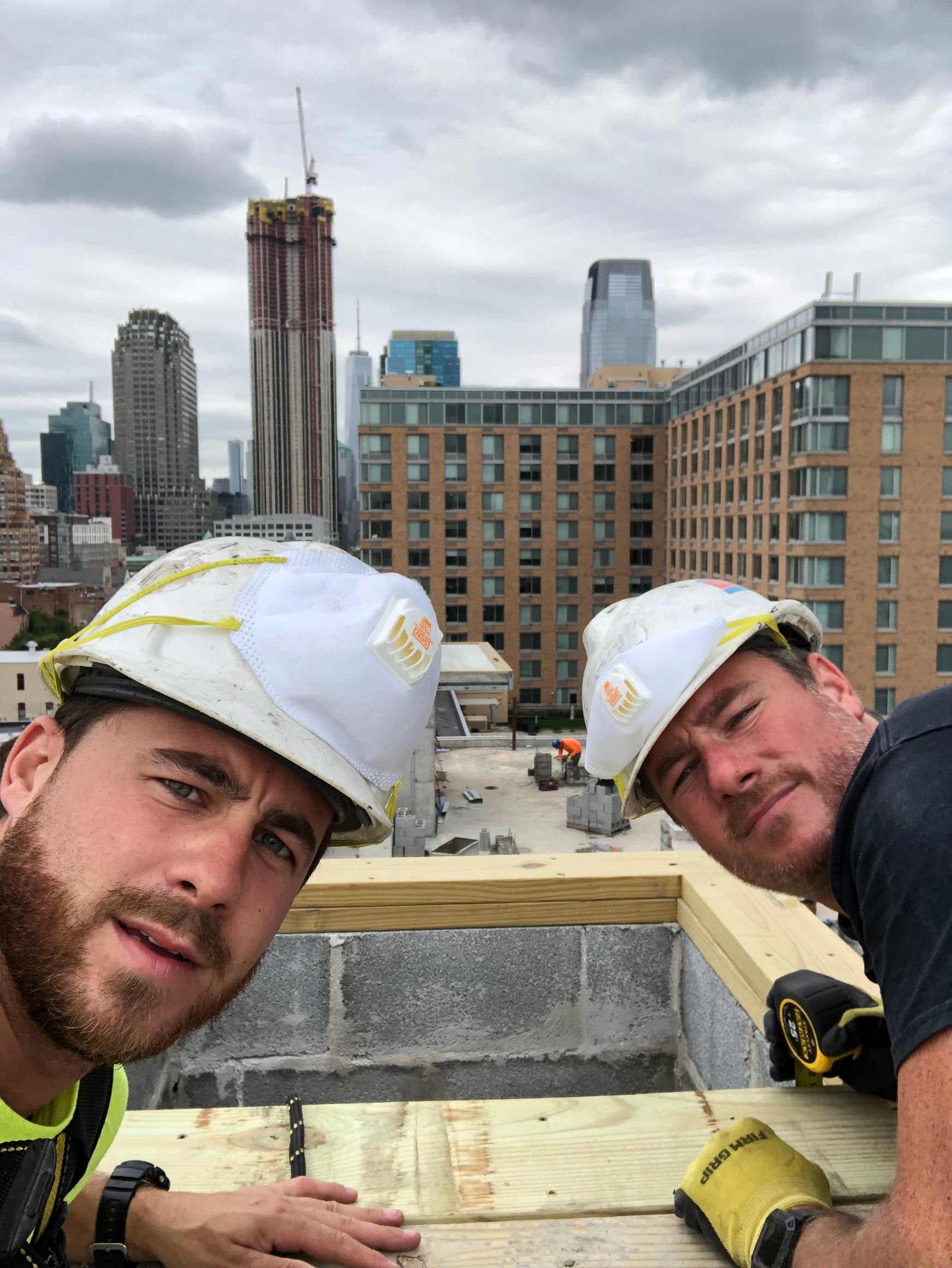 Two construction workers on a roof in a city, wearing helmets, looking at the camera.