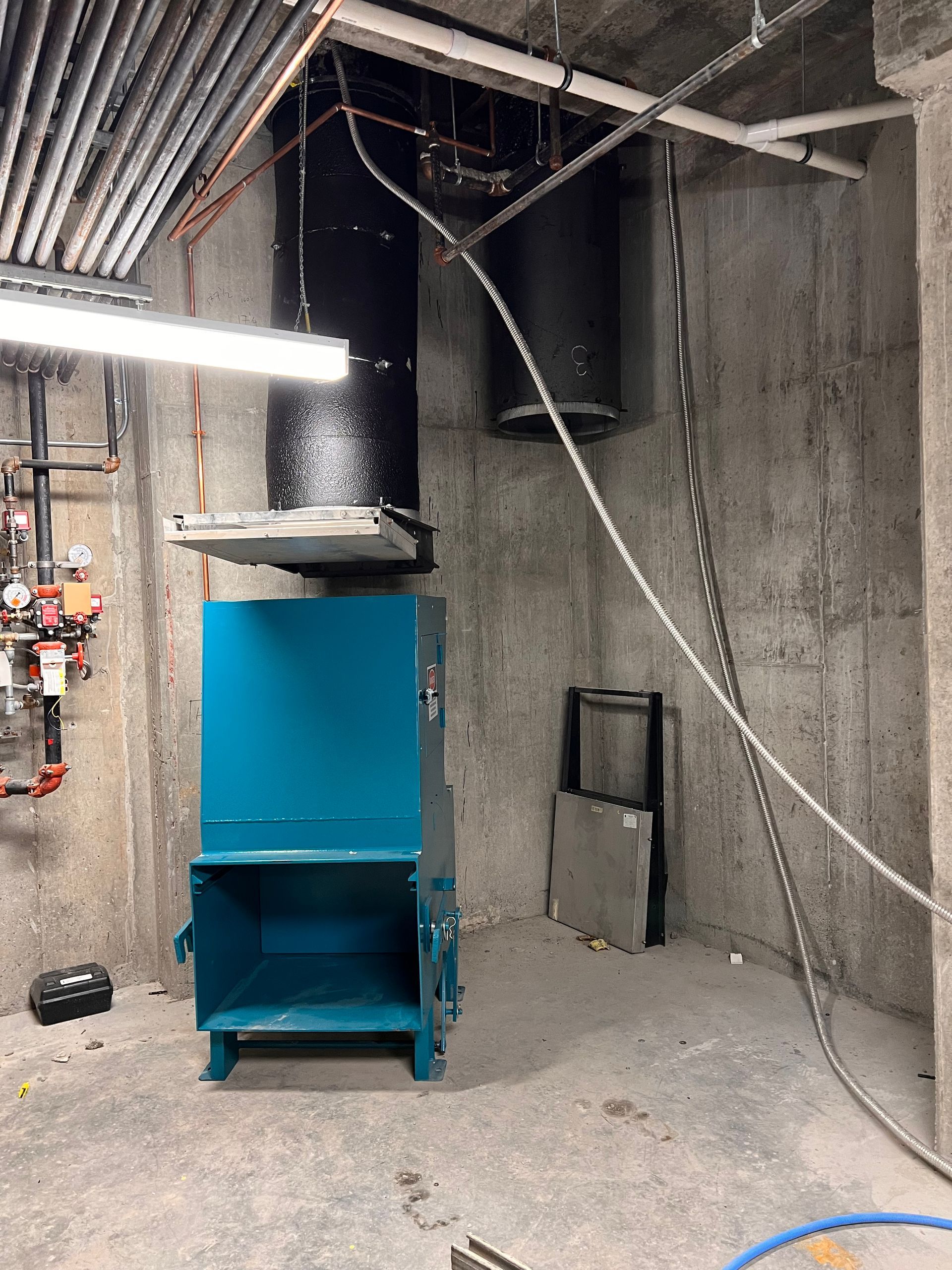 Blue trash compactor in a concrete utility room with ductwork overhead.