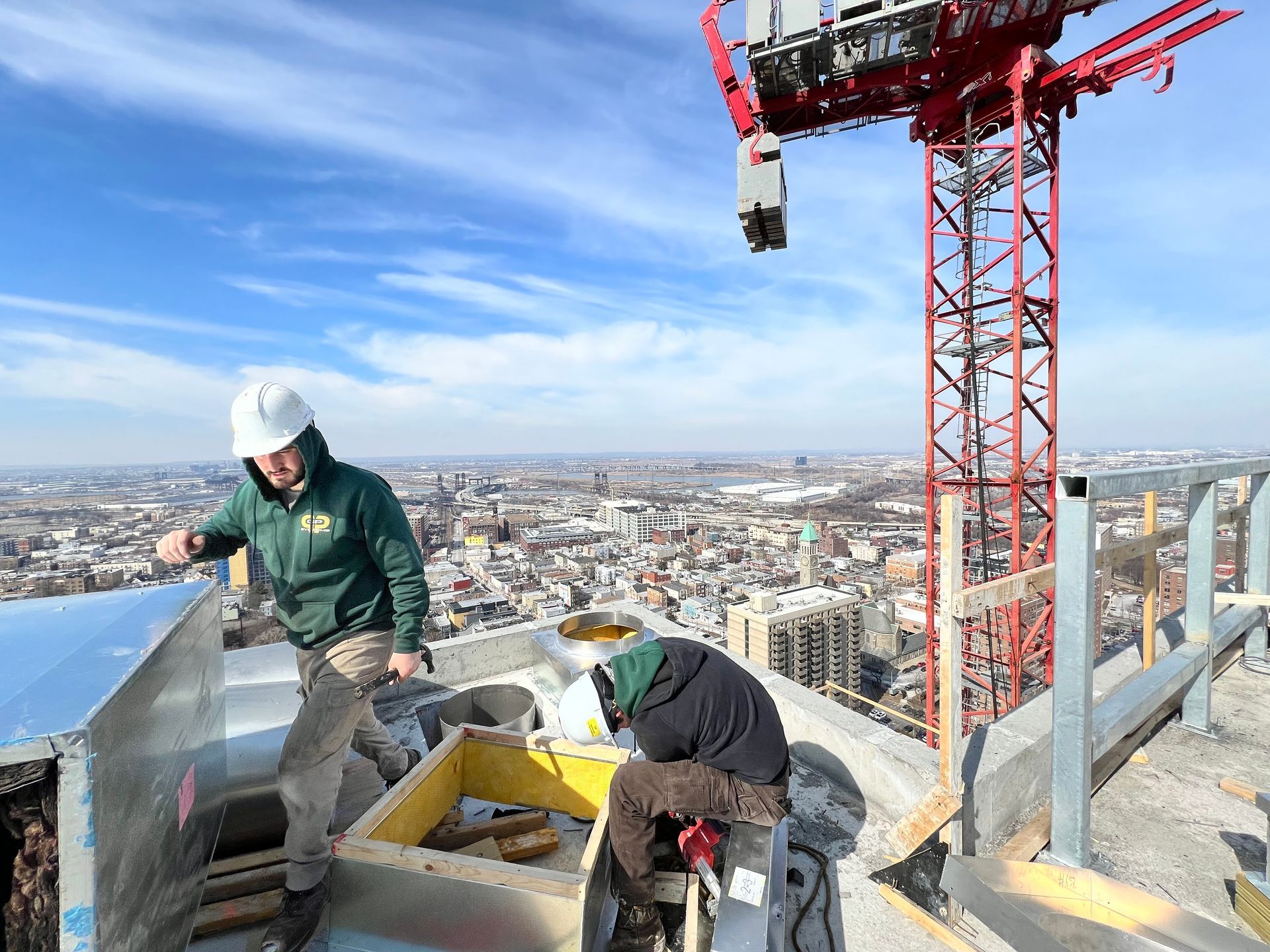 Two construction workers on a high-rise rooftop, installing equipment, with a city view and crane overhead.