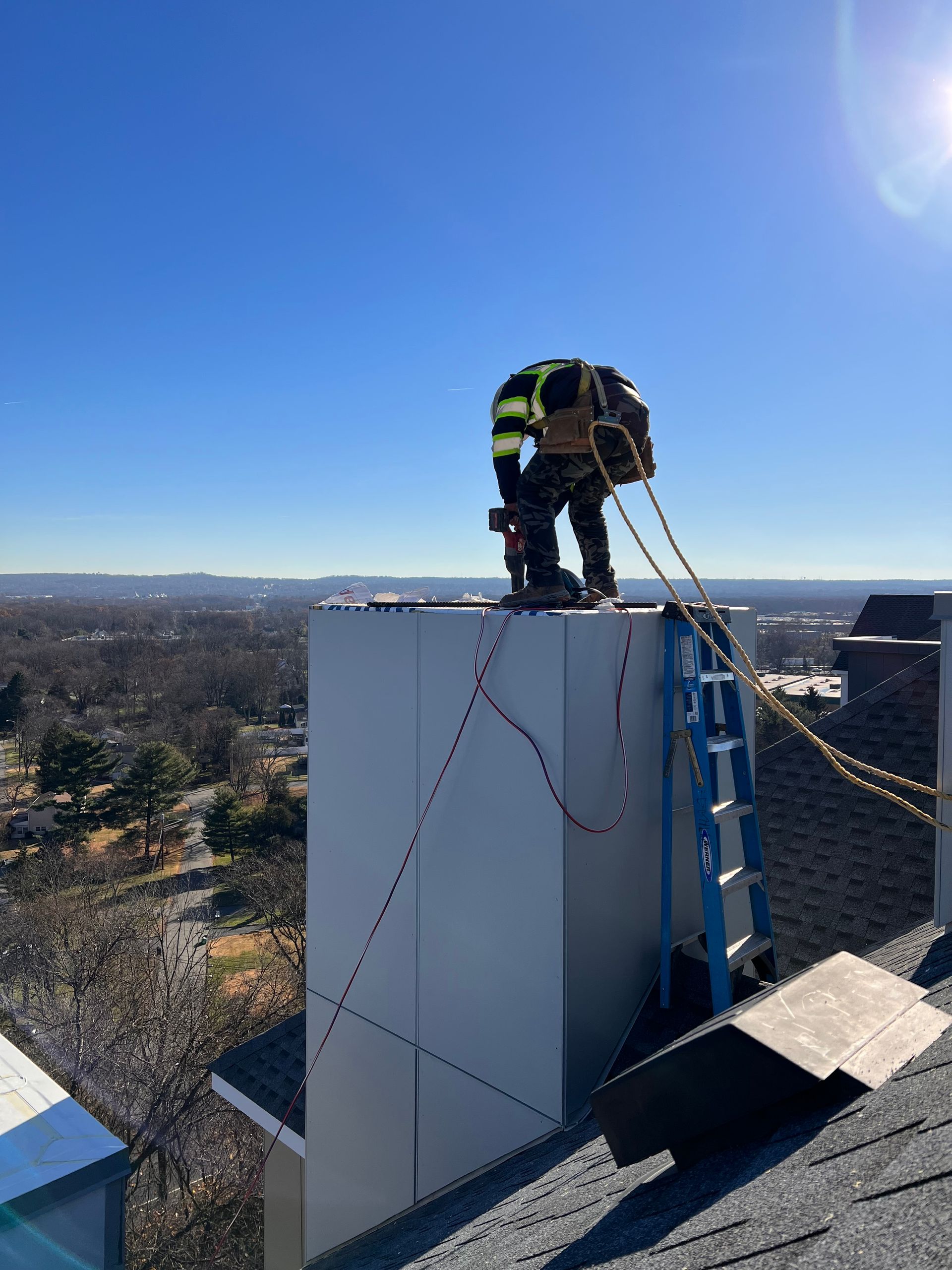 A worker in safety gear on a rooftop chimney. Ladder, ropes, blue sky, and cityscape visible.