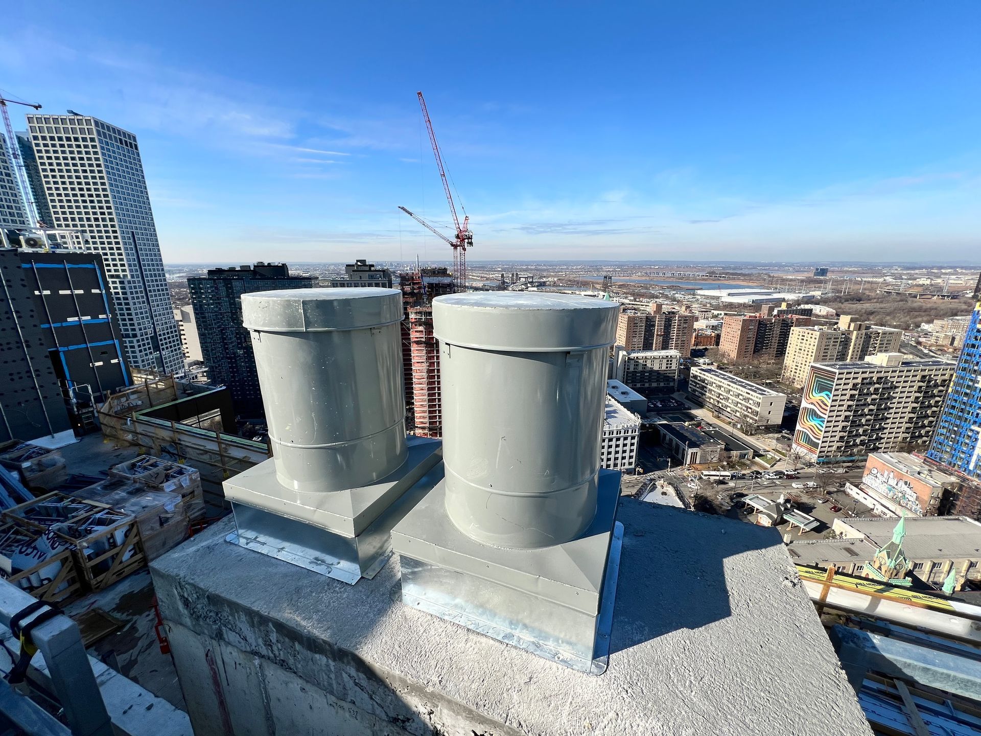 Two gray cylindrical rooftop vents with a city skyline in the background on a sunny day.