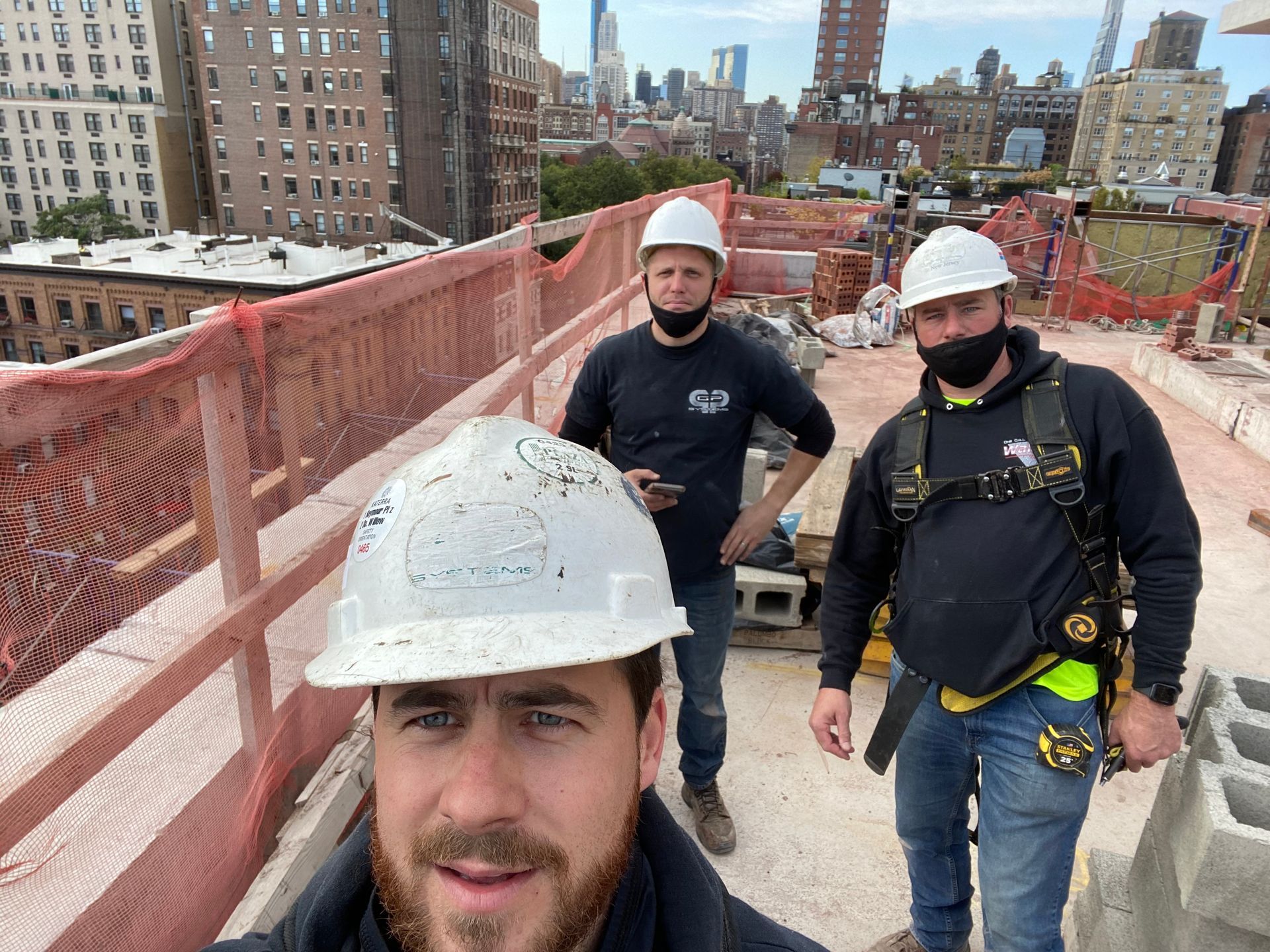 Three construction workers wearing hard hats and masks on a rooftop, with a cityscape in the background.
