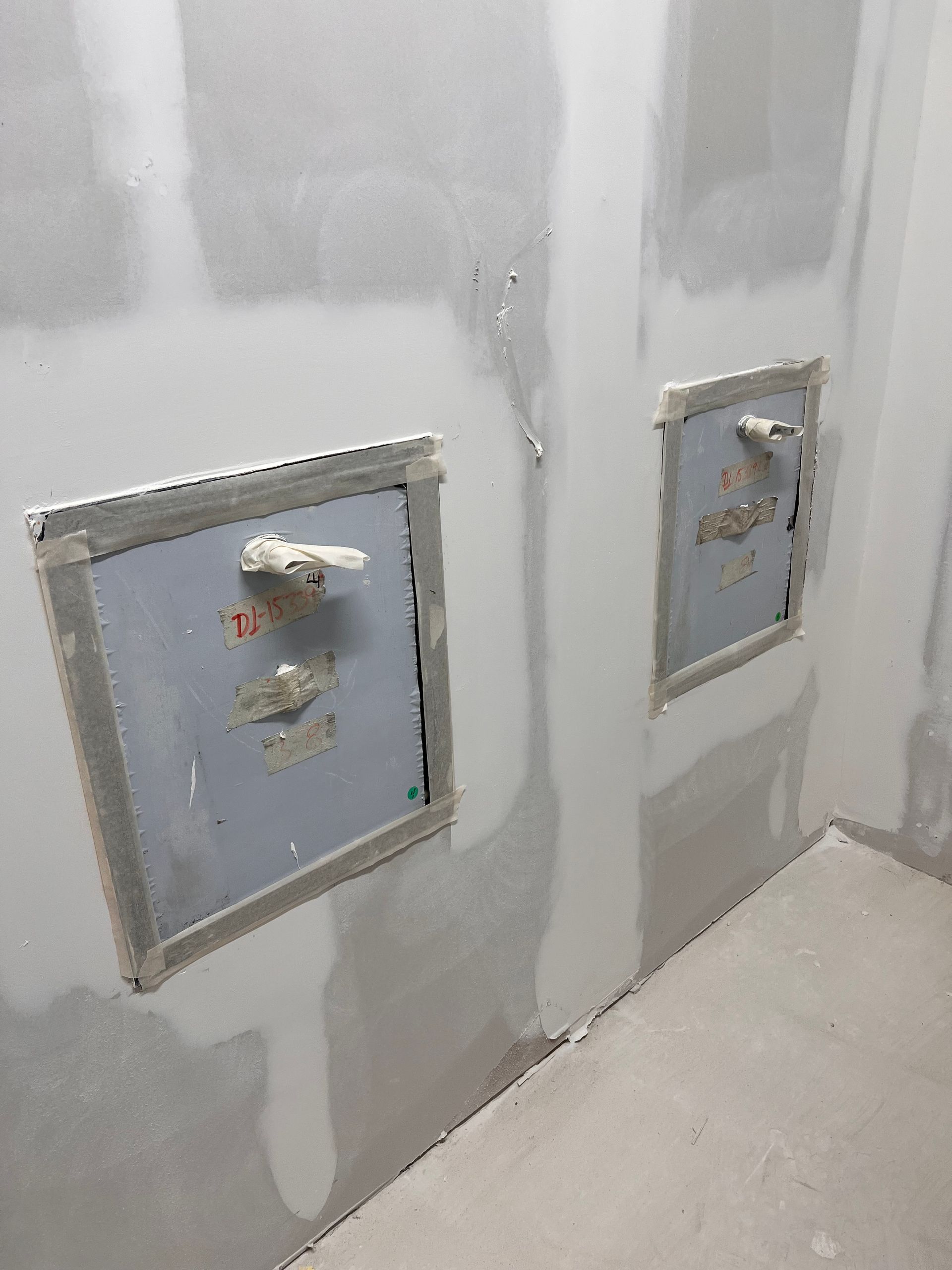 Two metal access panels in a drywall wall, with handles and gray sealant, in a room under construction.