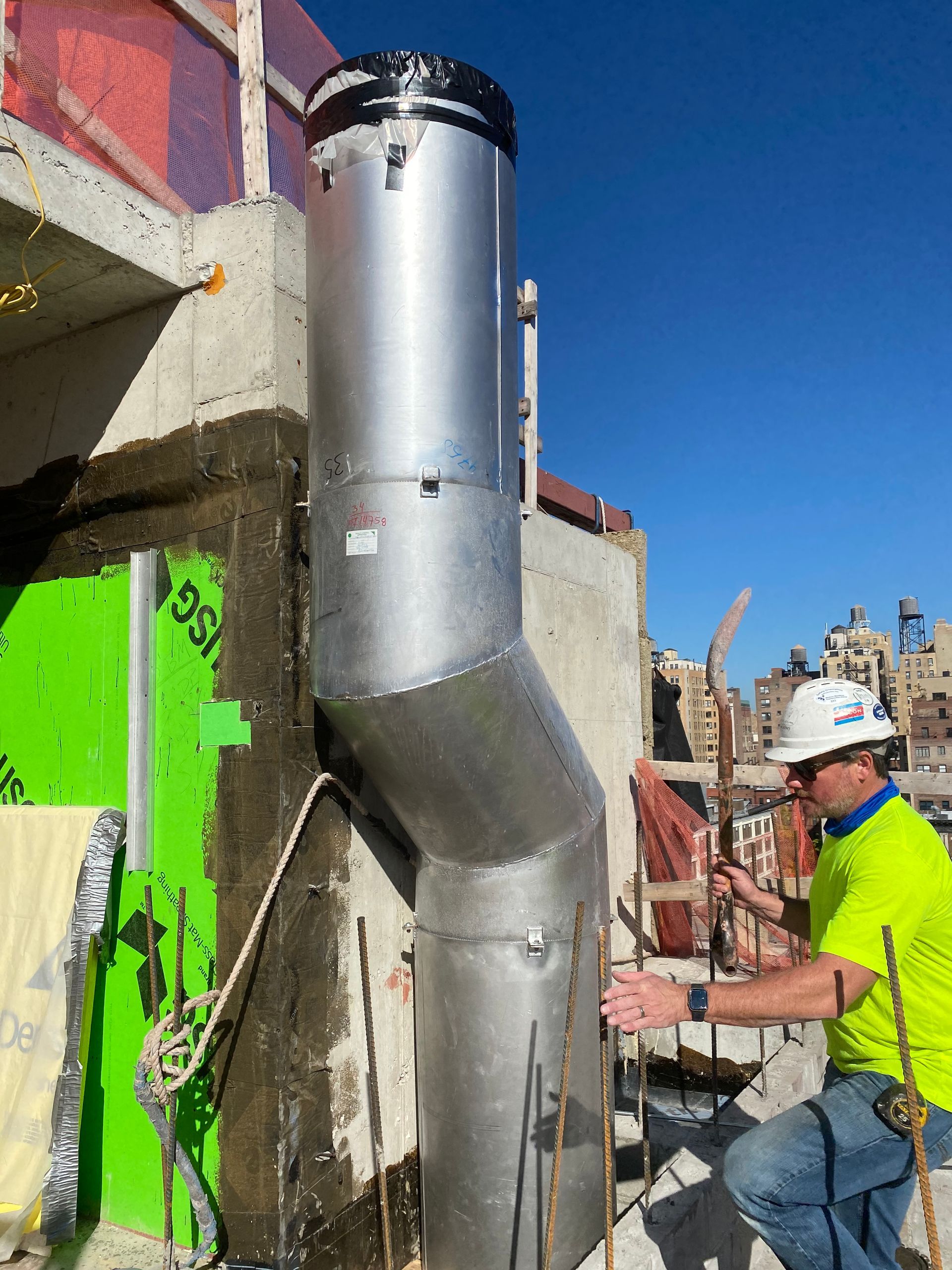 Construction worker inspecting a silver ventilation pipe on a building's exterior. Bright green insulation and blue sky.