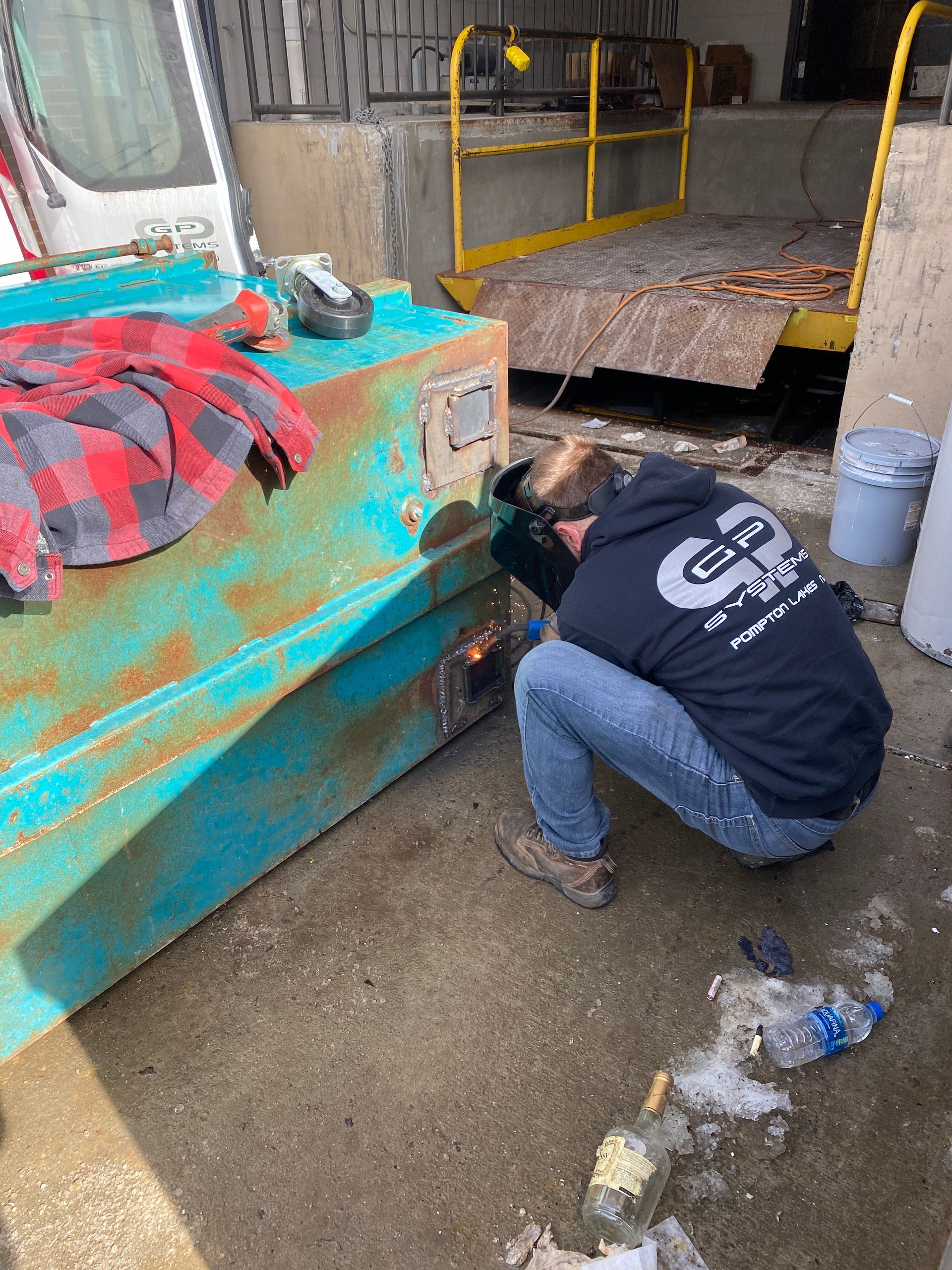 Person welds metal on a large blue tank, wearing a welding mask and jeans, outside.