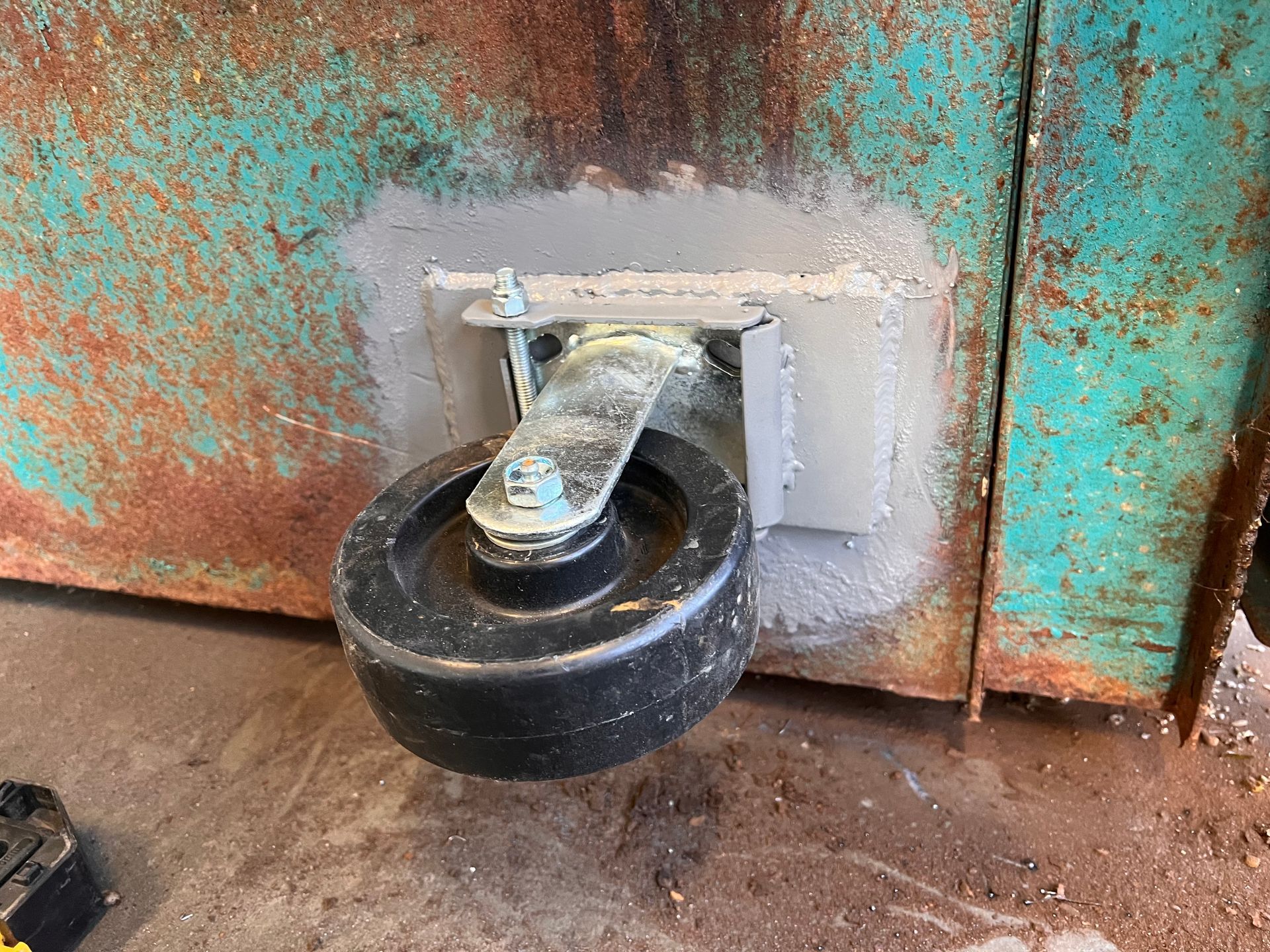 A metal wheel attached to a rusted, turquoise-painted surface, likely part of industrial equipment.