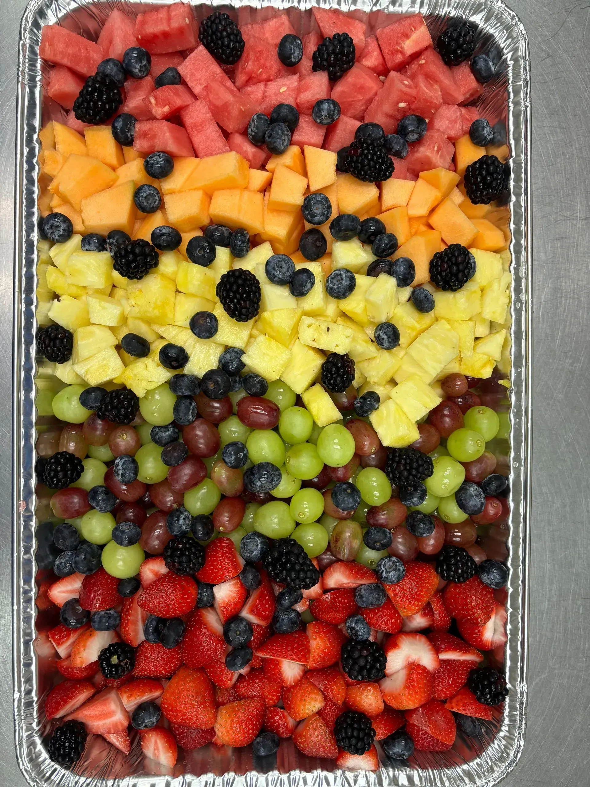Fruit tray arranged in rainbow order: watermelon, cantaloupe, pineapple, grapes, and strawberries. Blackberries and blueberries.