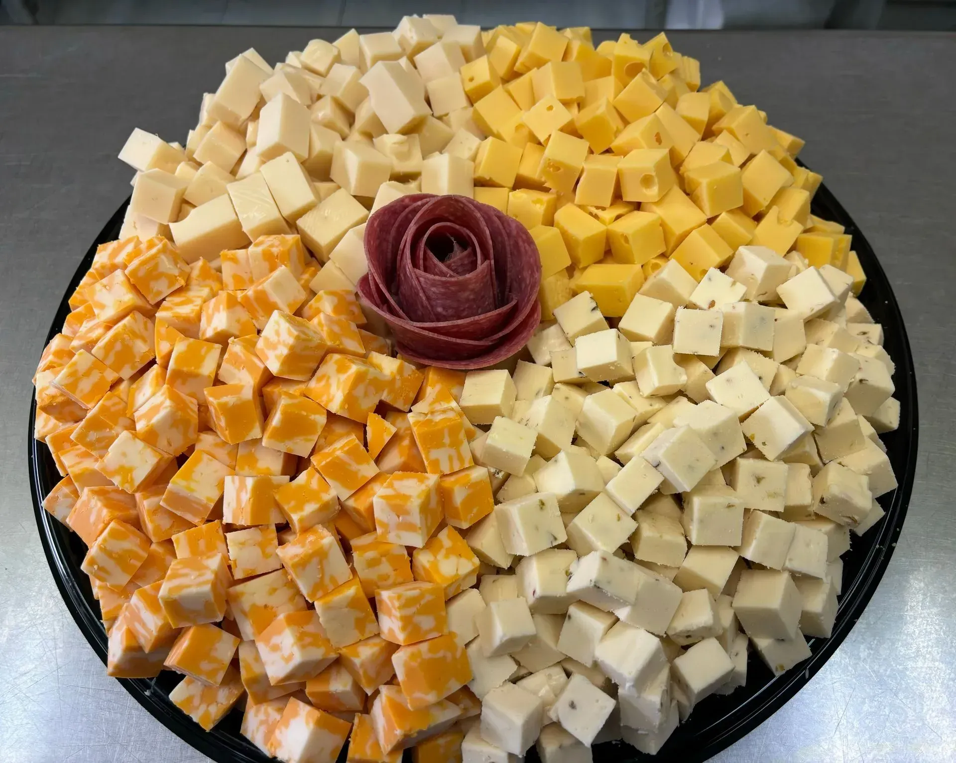A black platter with four types of cubed cheese arranged around a salami rose.