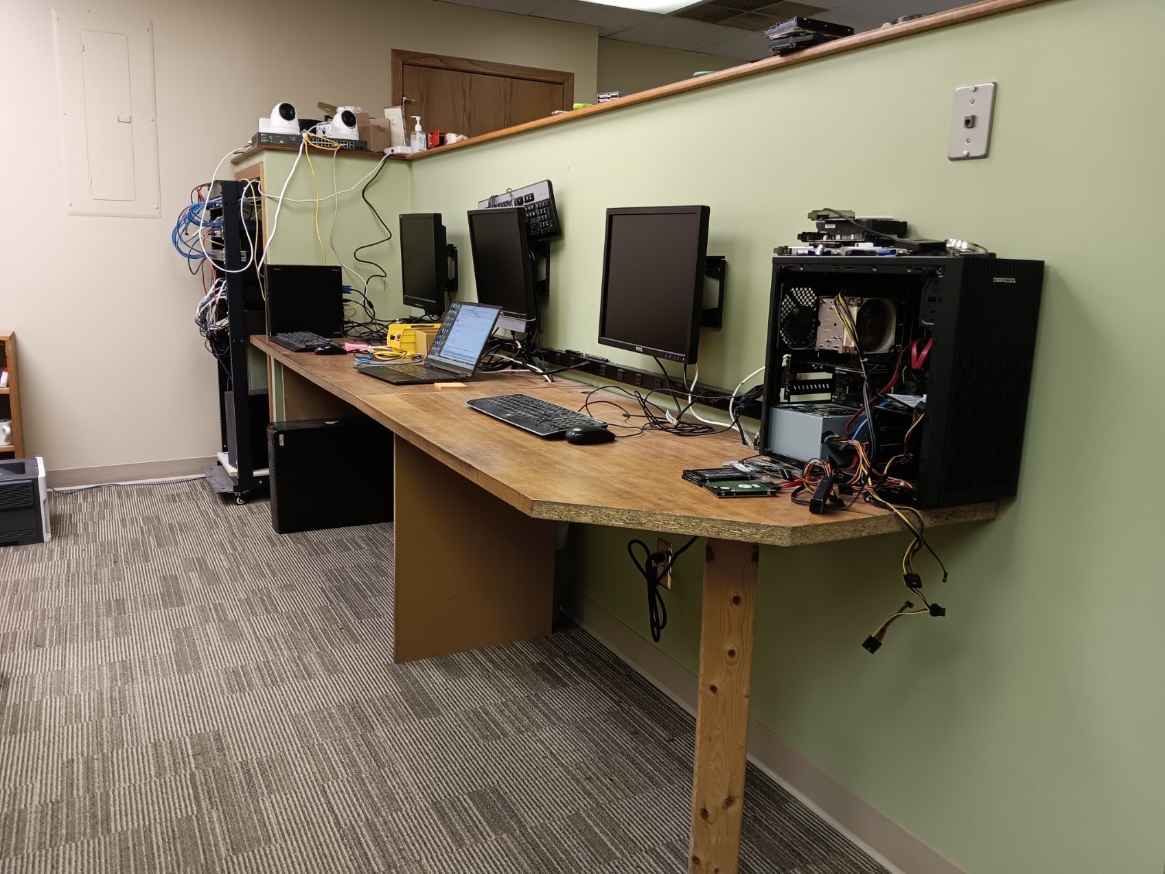 A workspace in an office features a long wooden desk with computer monitors, a tower case, and cabling against a wall.