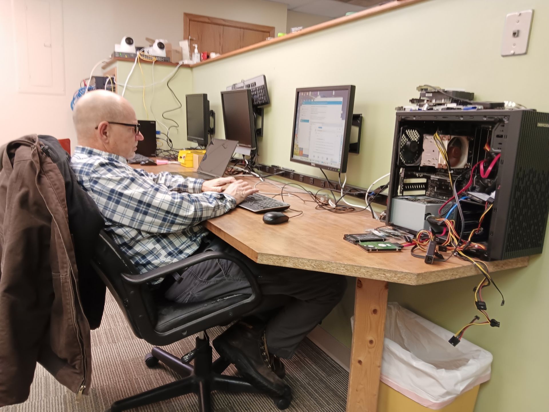A person works at a desk with an open computer tower and loose components, sitting in an office cubicle.