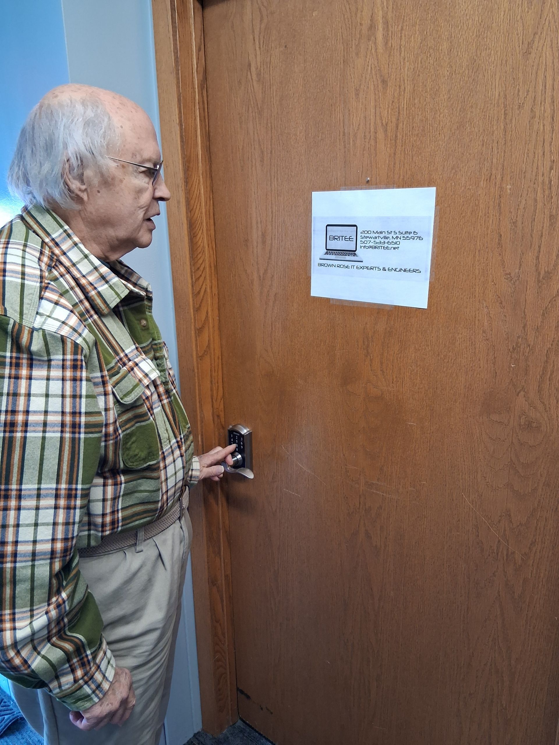 A person in a plaid shirt interacts with a digital keypad on a wooden door with a paper sign posted above the lock.