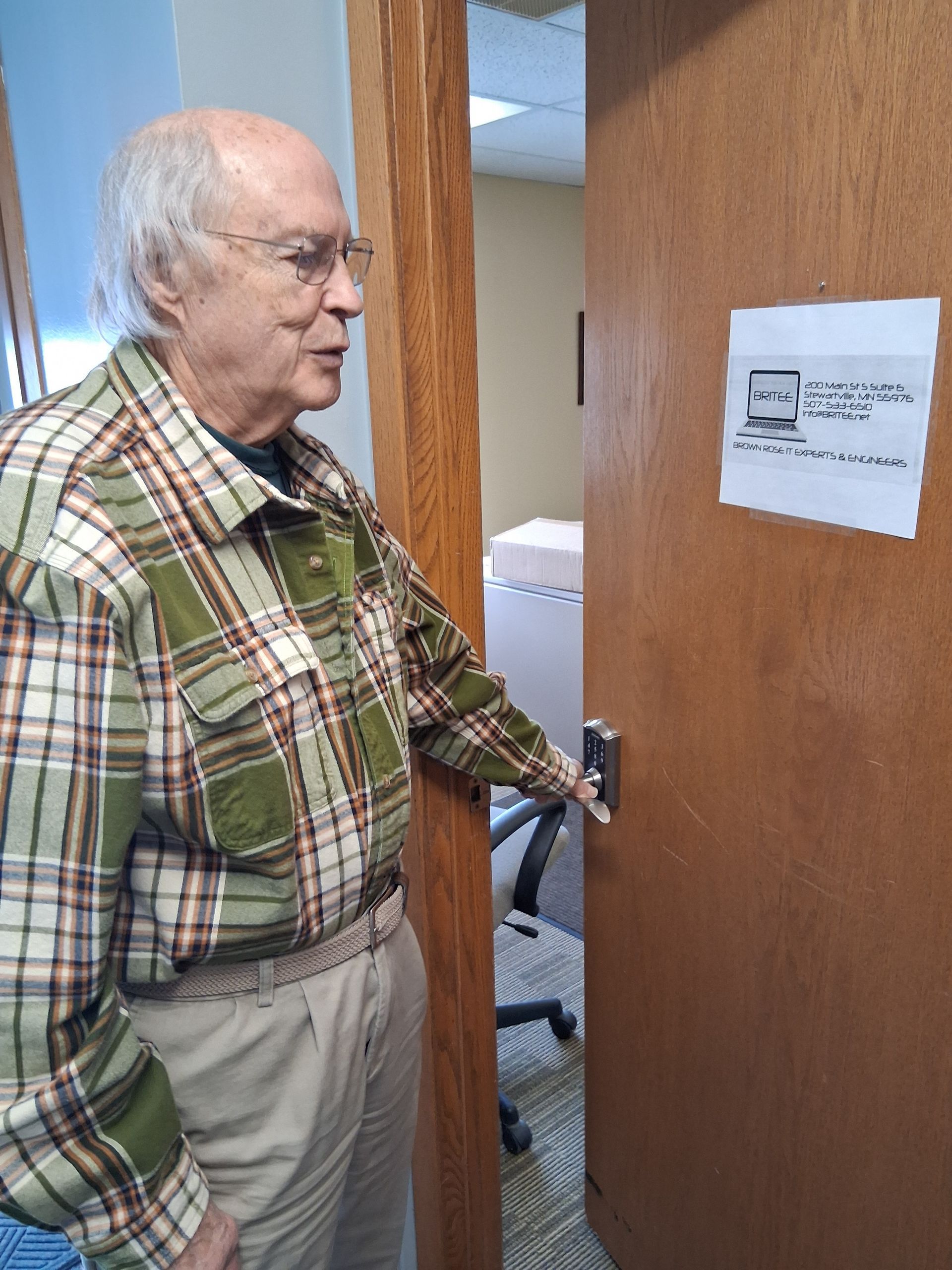 An elderly person wearing a green and brown plaid shirt opens a wooden office door with a computer icon sign on it.