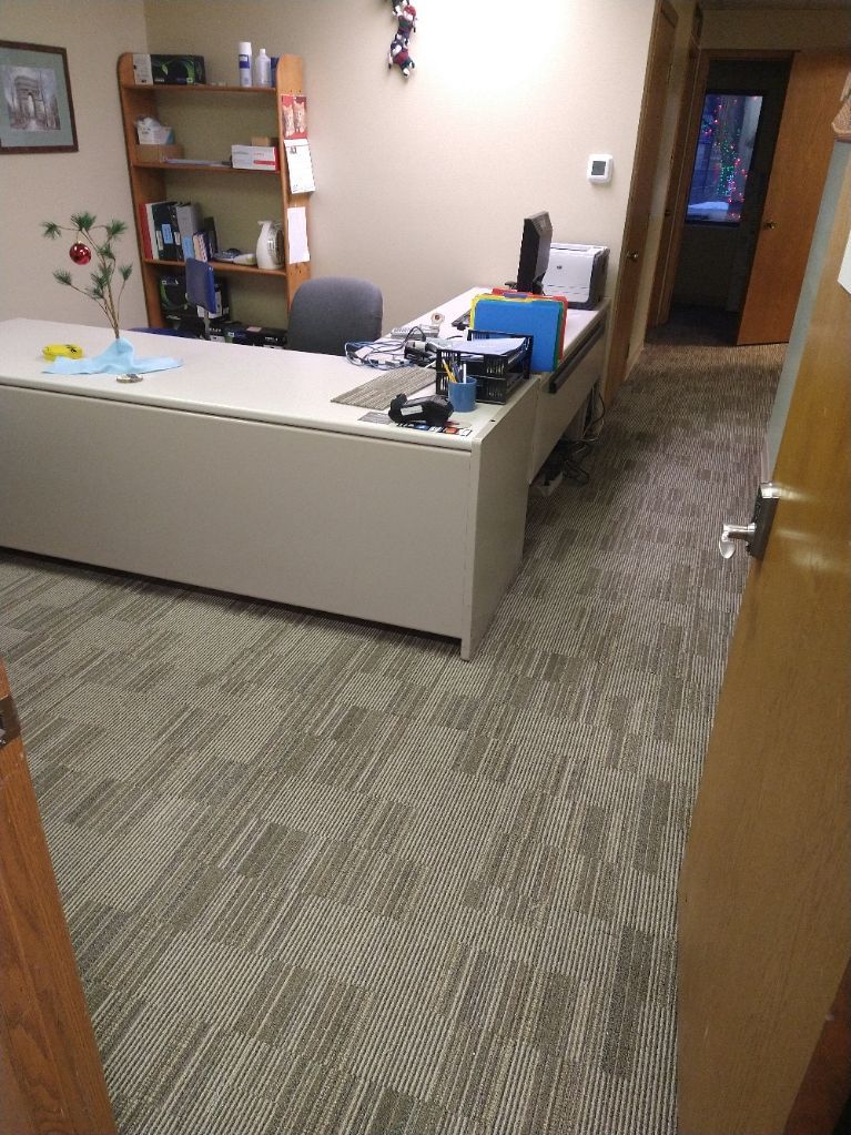 A view from a doorway into an office with a beige L-shaped desk, a wooden bookshelf, a computer, and patterned carpet.