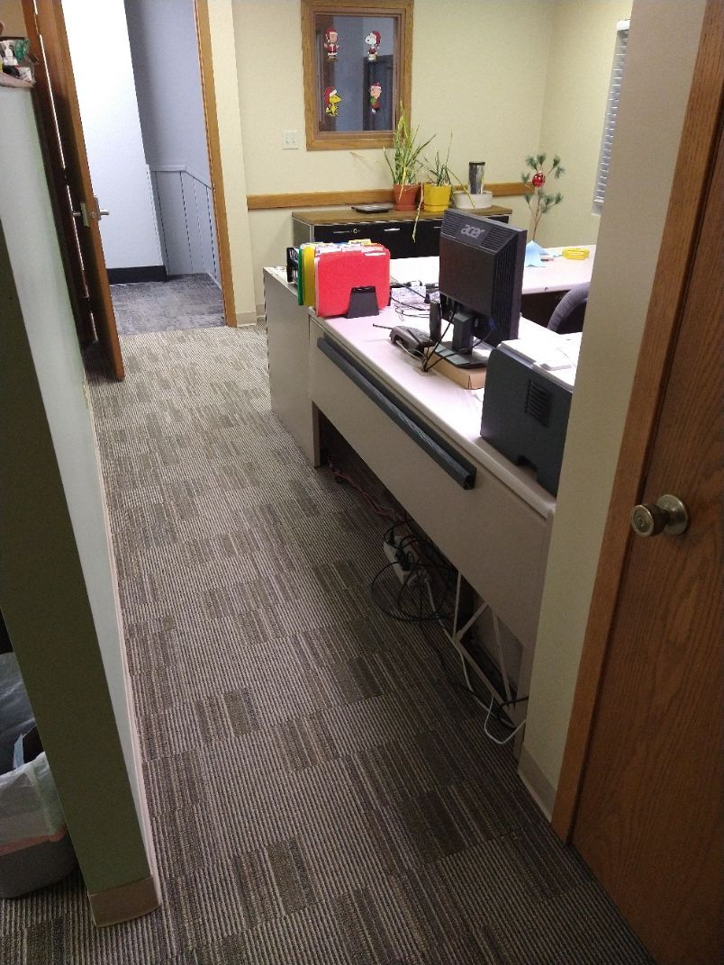A narrow office hallway leads to a desk with a computer monitor, colored file folders, and a plant against a wall.