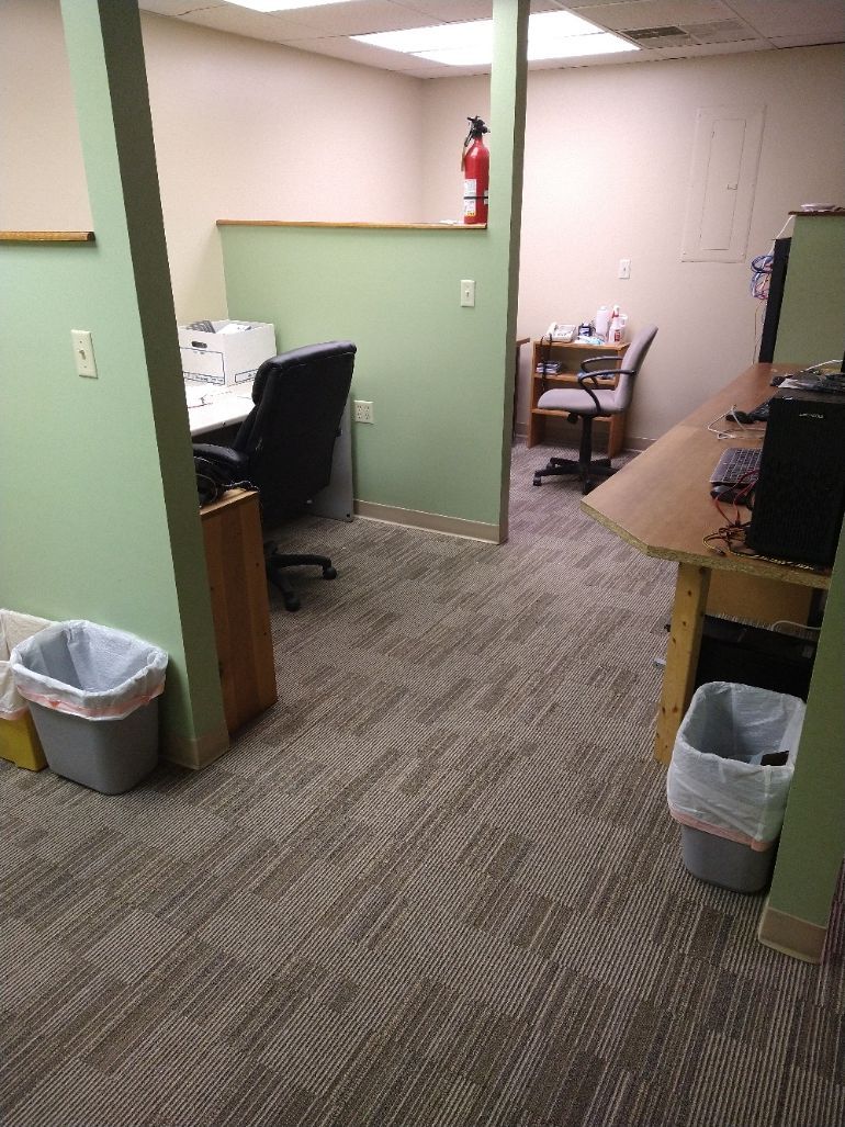 Office space with patterned carpet, two desks, black office chairs, a fire extinguisher, and trash cans.