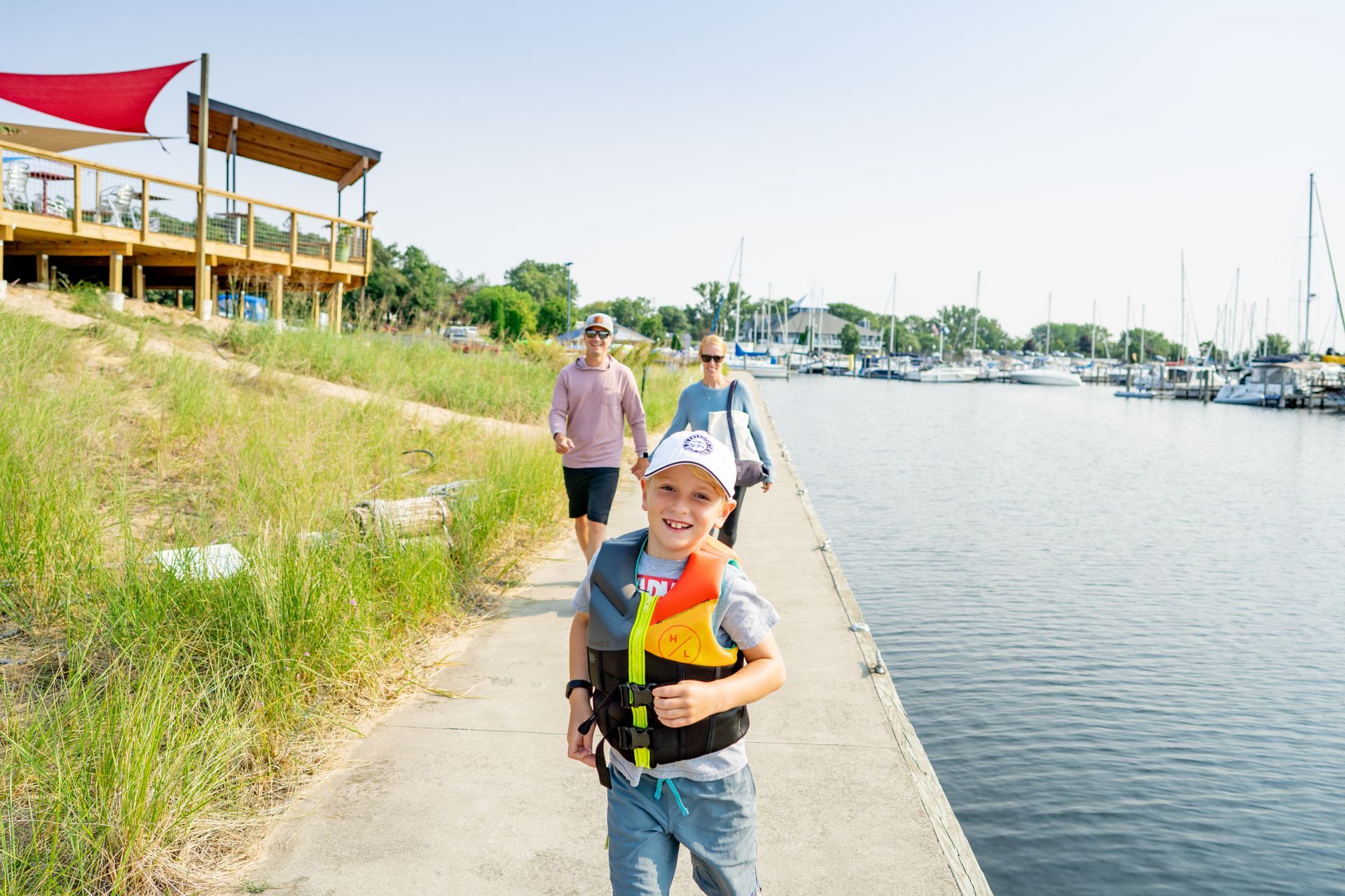 Boy in life vest runs towards the camera on a dock, with parents walking behind, marina and building in the background.