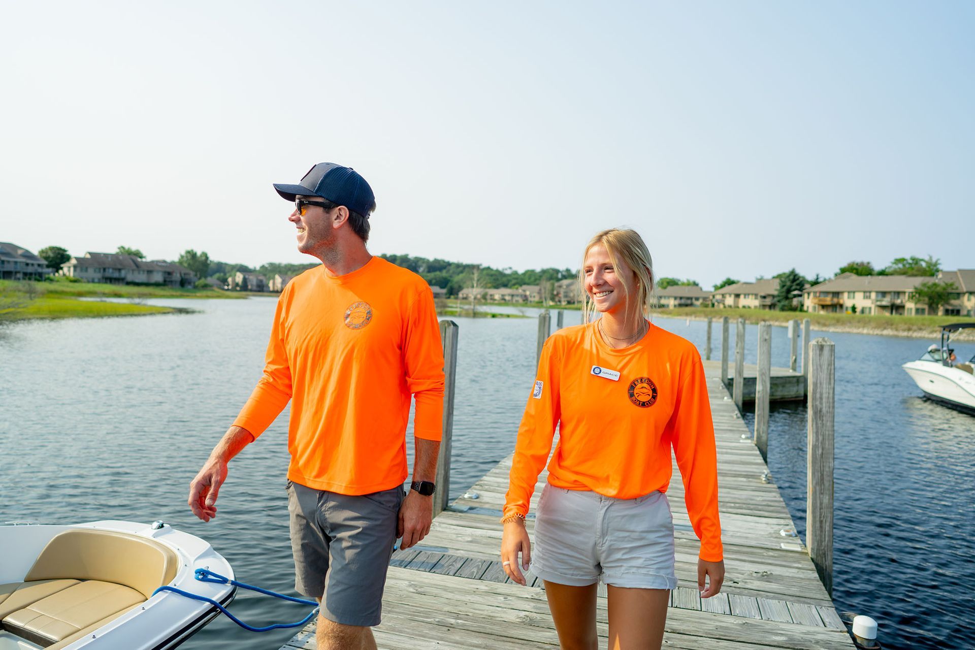 Two people in orange shirts walk on a wooden dock on a sunny day, looking at the water.