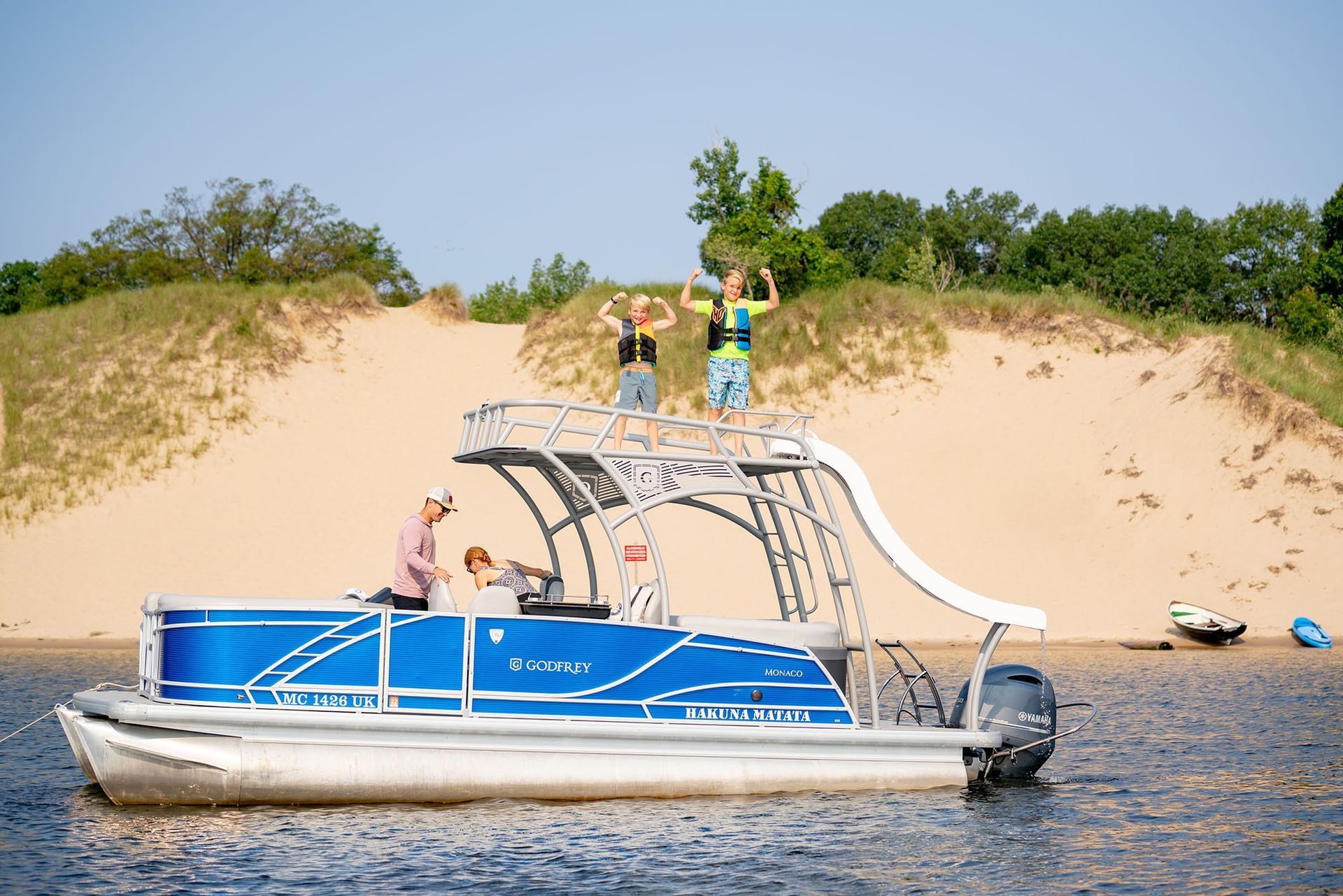 Pontoon boat with slide, two people on top, others seated. Blue boat on water near dunes.