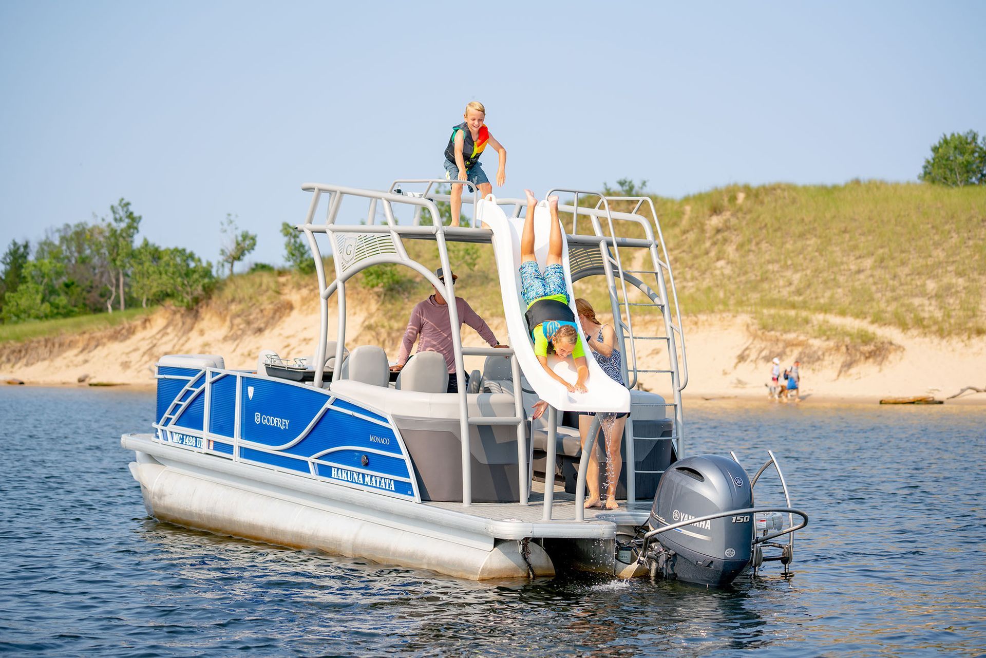 Pontoon boat with slide. People playing; one sliding into water. Blue and silver boat on lake near sand dunes.