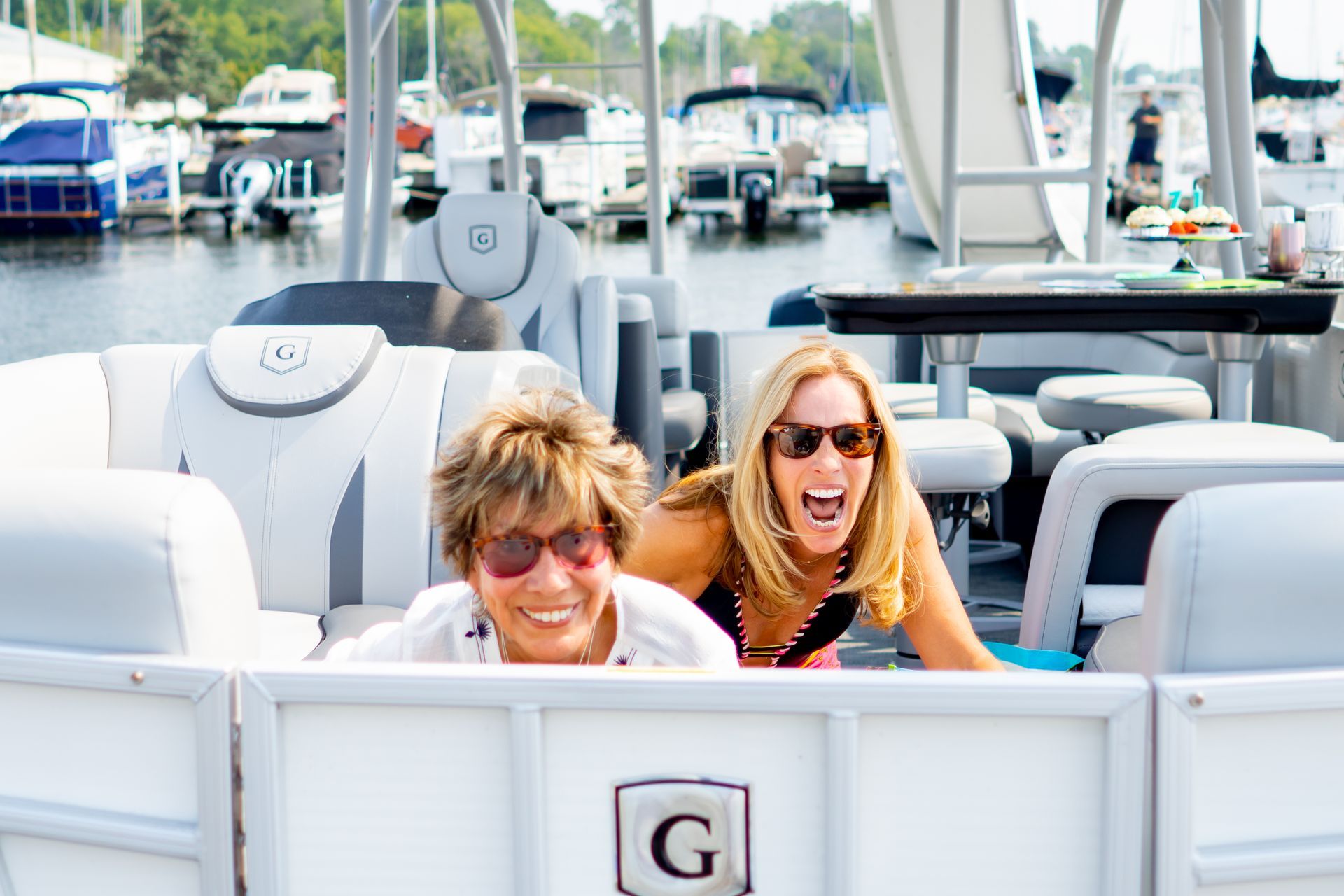 Two women on a pontoon boat, smiling and laughing. Harbor and other boats in background.