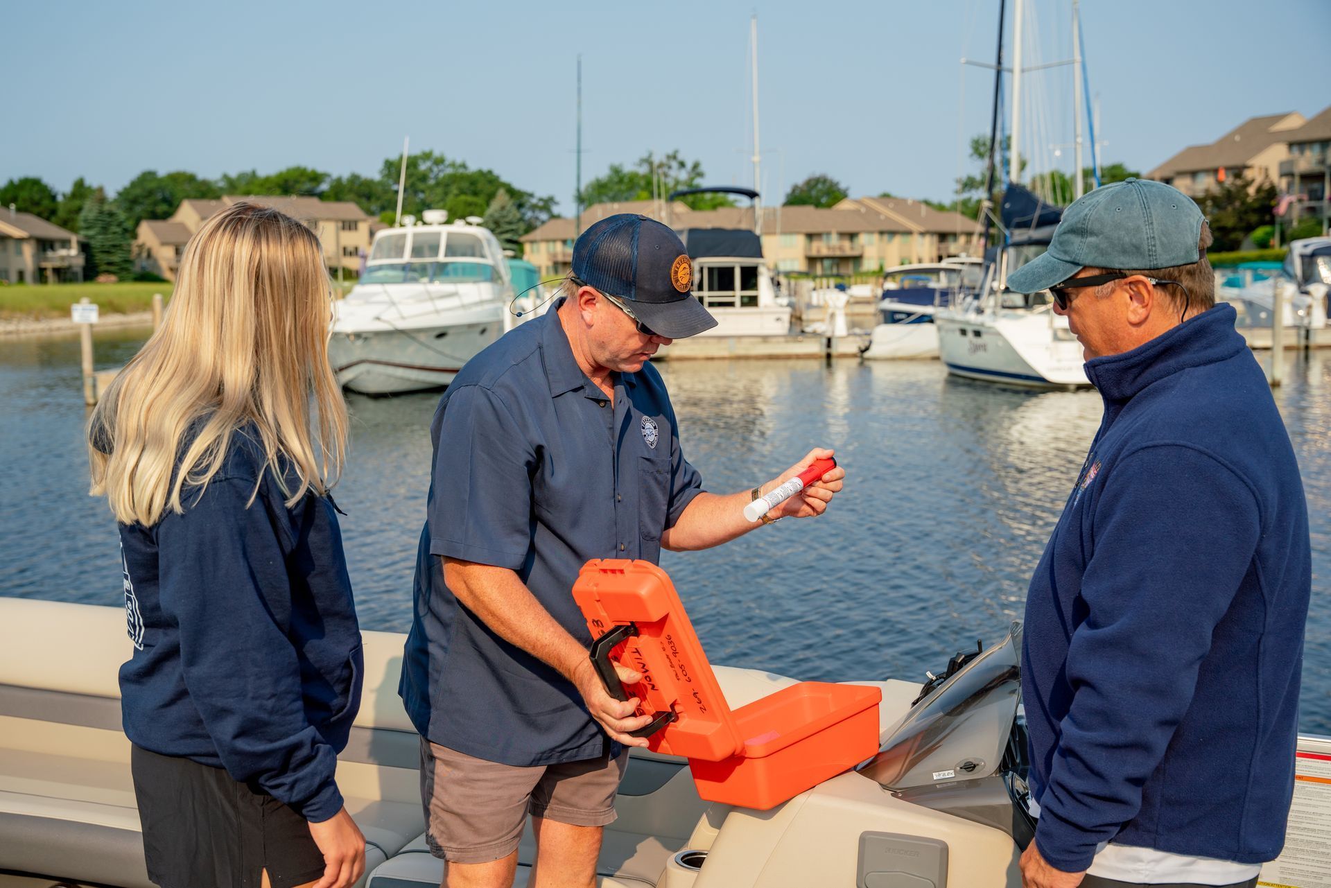 Three people at a marina; man holds orange emergency kit, demonstrating its use. Boats and water in background.