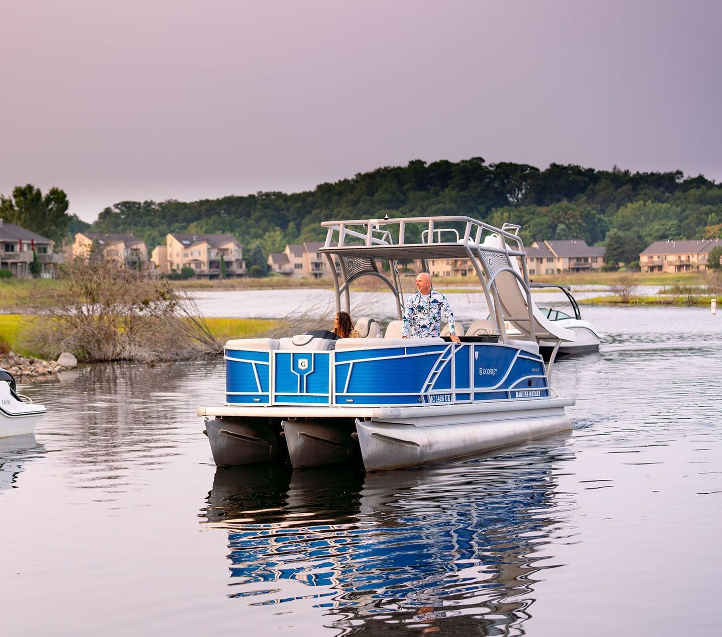 Blue pontoon boat on water, two people aboard, houses and trees in the background.