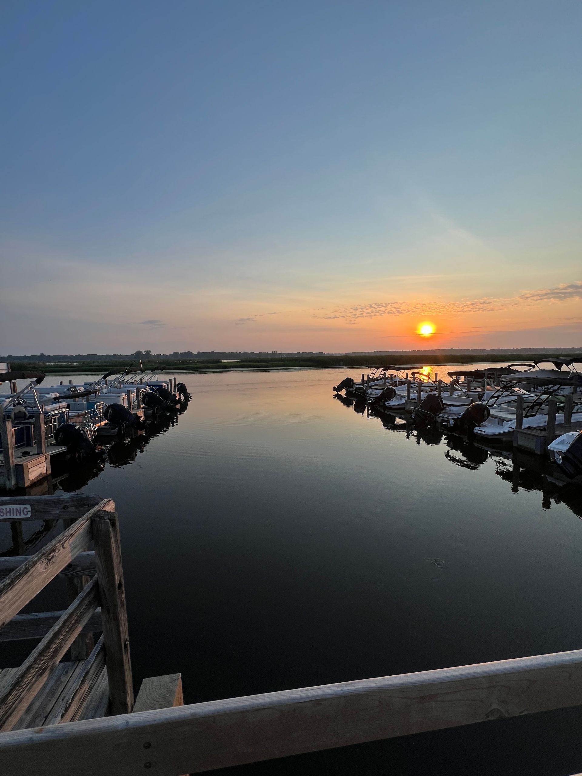 Sunset over a marina with boats docked, calm water reflecting the orange sun and blue sky.