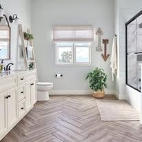 Bright modern bathroom with white vanity, toilet, window, plant, and glass shower on herringbone tile floor