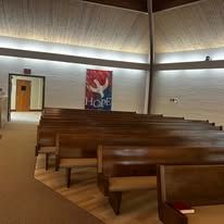 Empty church sanctuary with wooden pews, slanted ceiling, and a colorful wall banner near the front.
