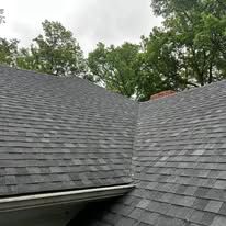 Gray shingled roof with a steep valley, viewed against leafy green trees and cloudy sky