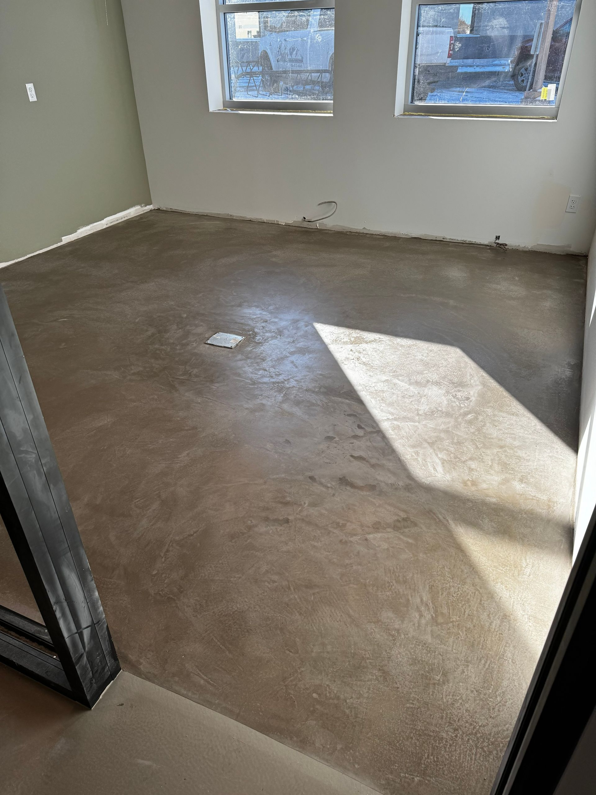 Empty room with unfinished concrete floor, white walls, and sunlight from two windows.