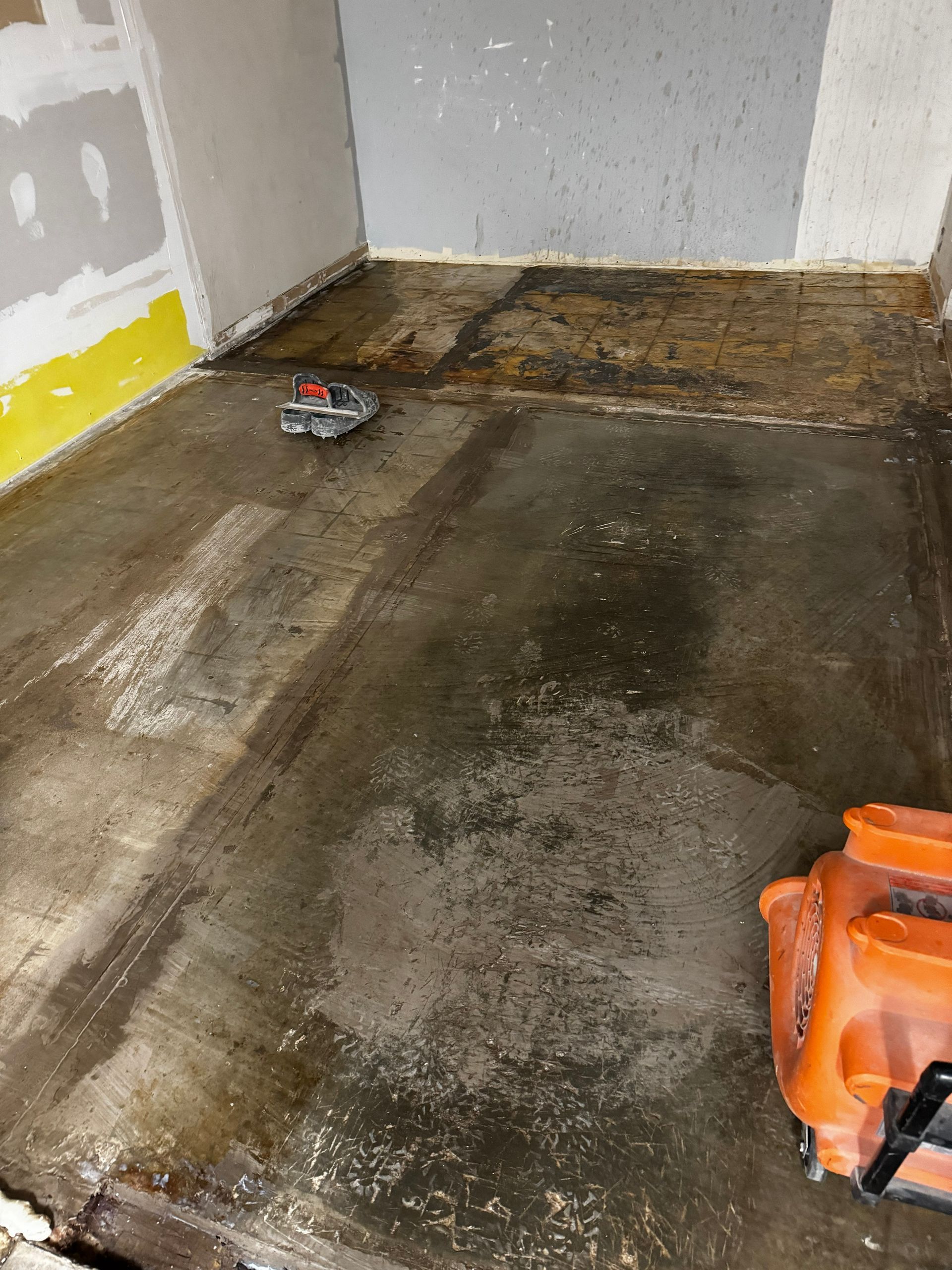 Damp, stained room corner with dark floor, peeling walls, and an orange tool at lower right.