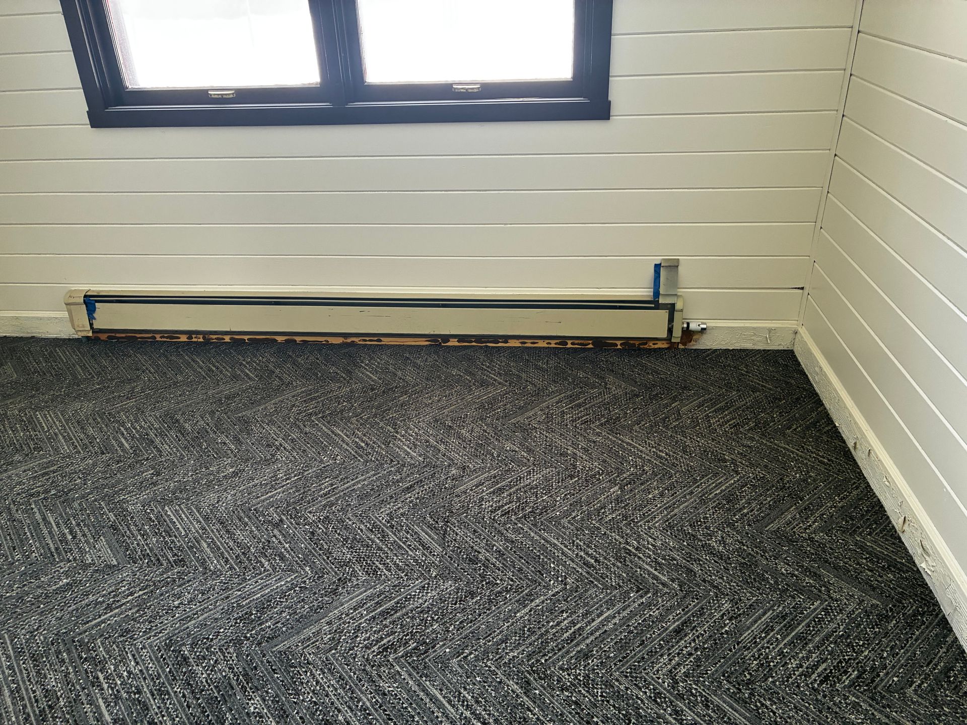 Empty room corner with window, white paneled walls, and dark patterned carpet.